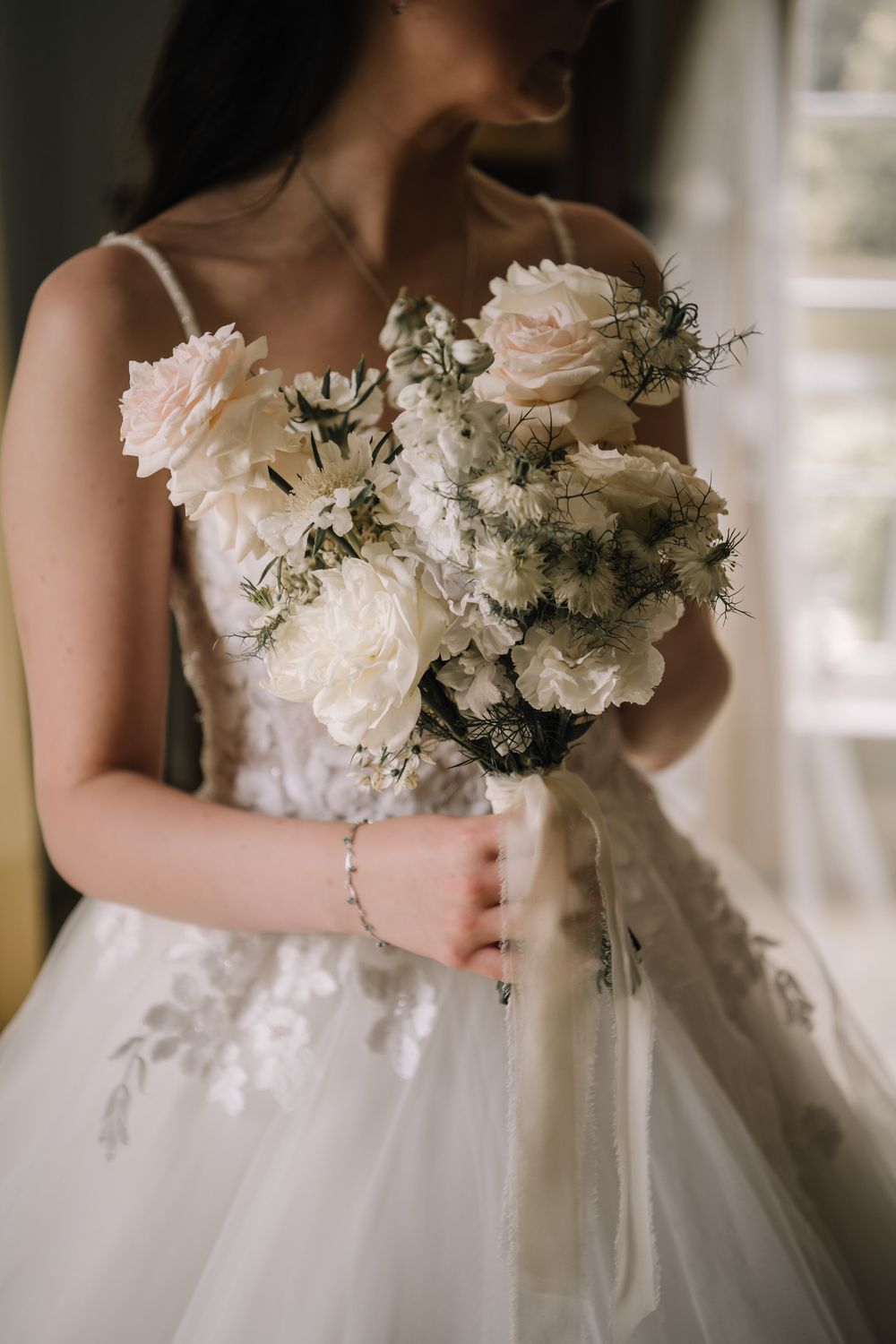 Bride in lace wedding gown holding elegant white and blush floral bouquet with roses and wildflowers near a window.