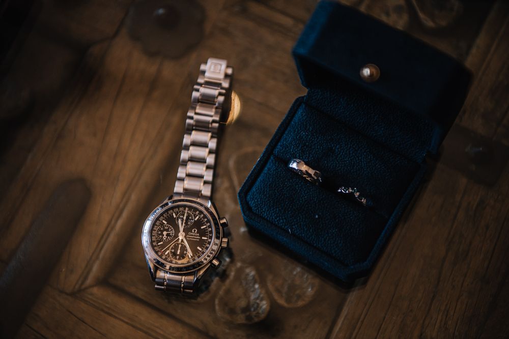Silver chronograph watch beside a blue velvet ring box with wedding bands on a wooden surface.
