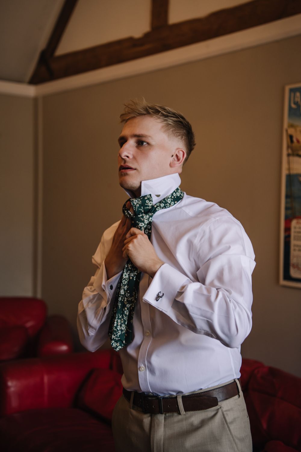 A young groom in a white shirt adjusts his green floral tie while getting ready indoors before his wedding ceremony.