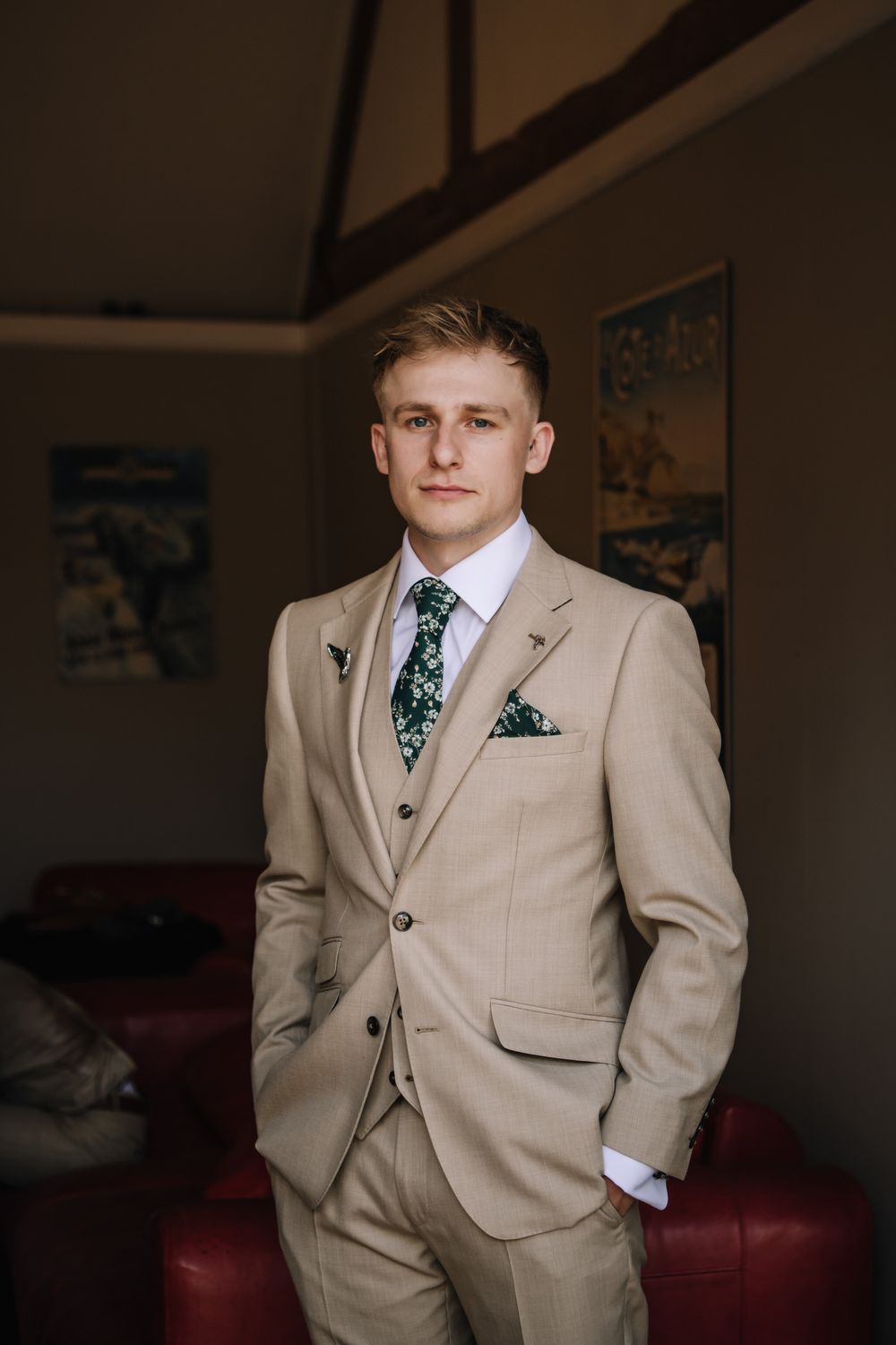 Young groom in beige three-piece suit with green floral tie and pocket square stands confidently indoors.