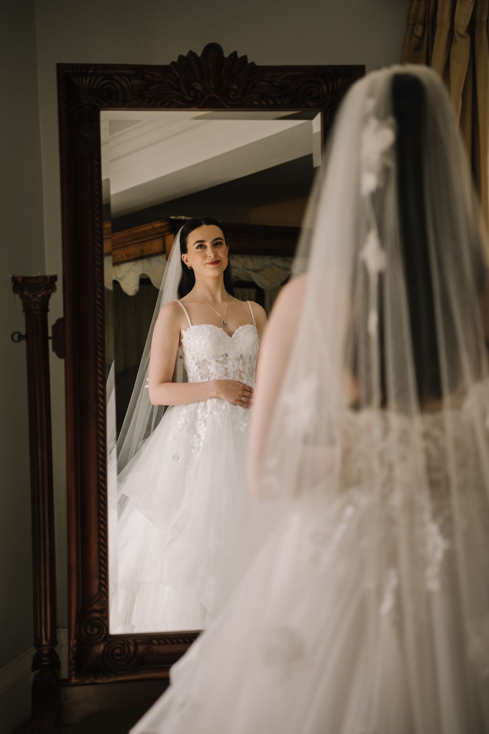 Bride in white lace wedding gown and veil admiring her reflection in an ornate dark wood mirror.