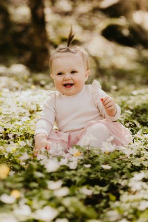 Baby wearing pink sits among blooming white flowers in a dreamy outdoor setting.