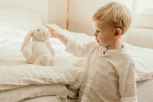 A toddler in white pajamas sits on a bed reaching toward a plush bunny toy in soft morning light.