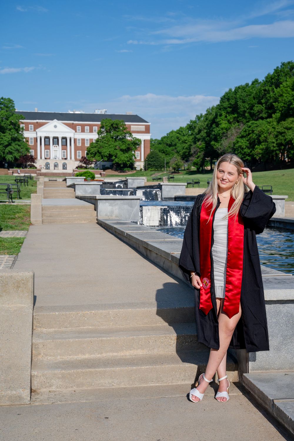 College graduate in her gown poses on steps near fountain with McKeldin Library and blue sky in background.