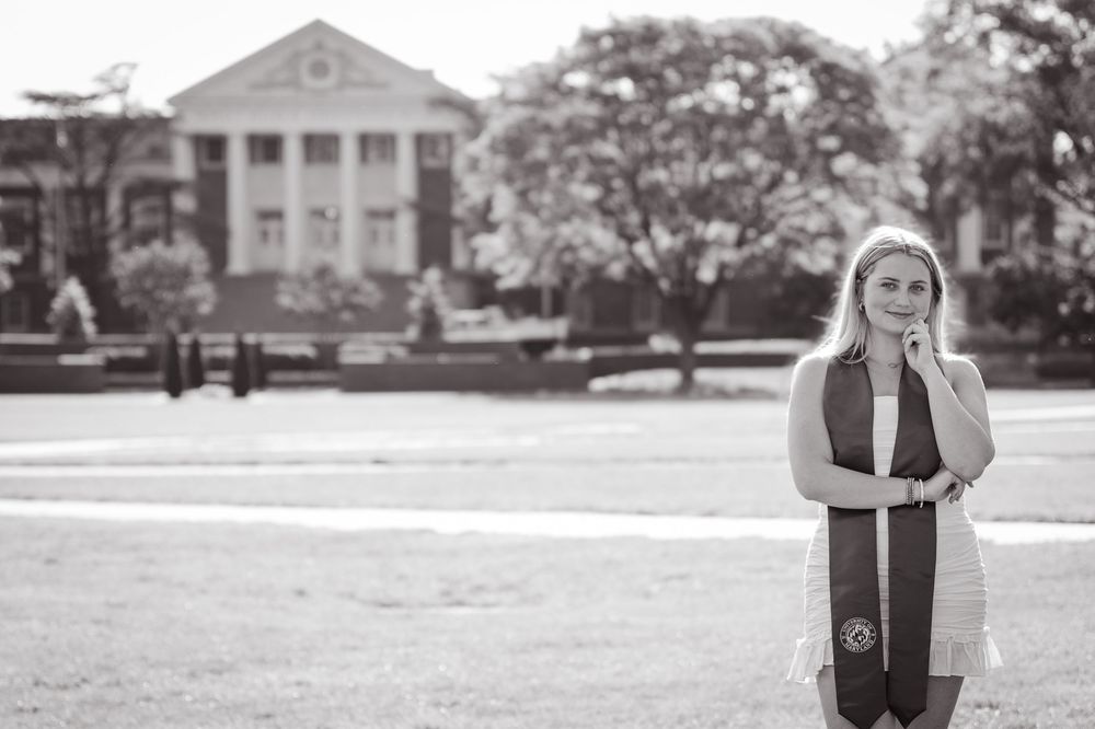 A student in graduation attire stands on UMD's college campus lawn with a grand white-columned building in the background.