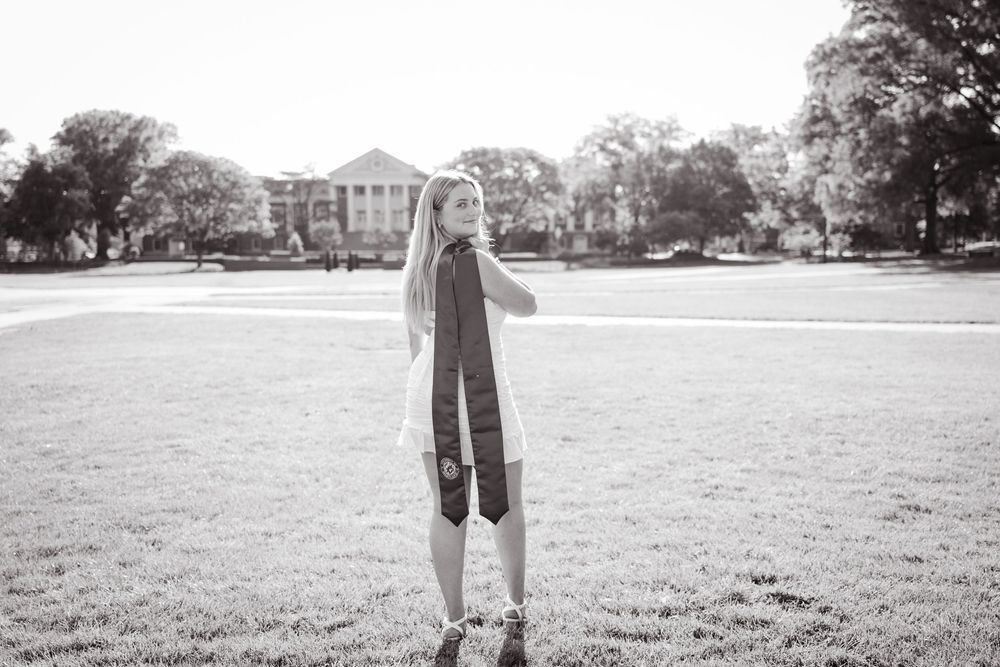 Black and white portrait shows a college graduate on a grassy lawn with a campus building visible in background.