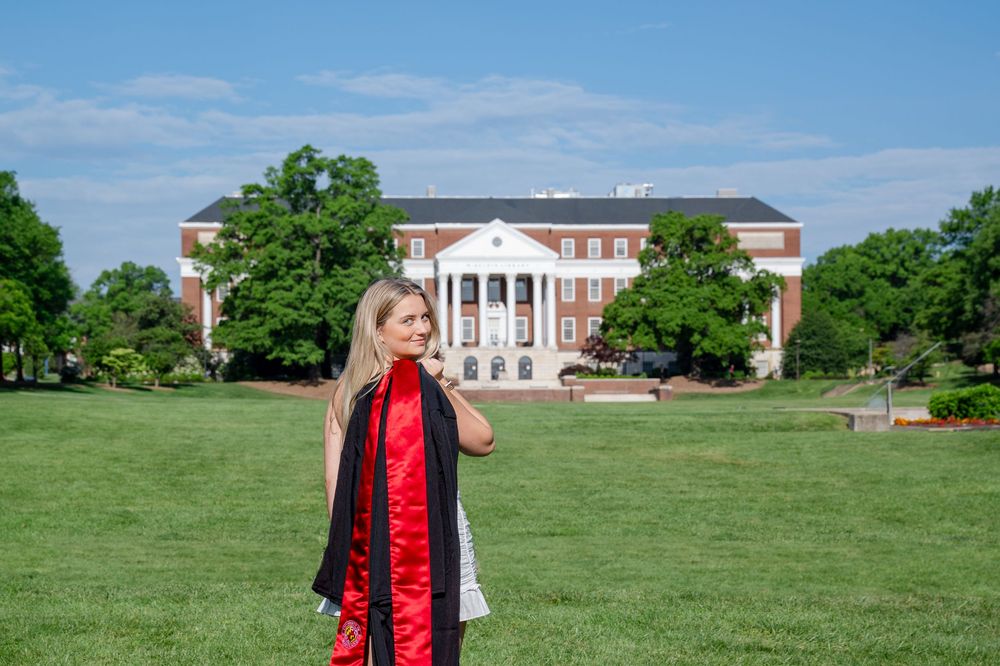 A college graduate stands on the green lawn in front of McKeldin Library on UMD's campus.