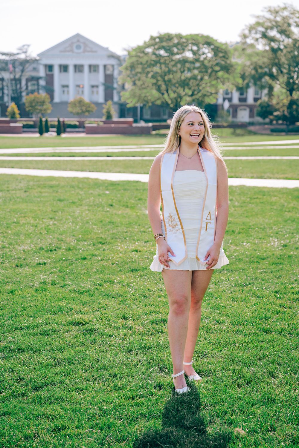A college graduate in a white dress stands on the campus lawn with a stately columned building in the background.