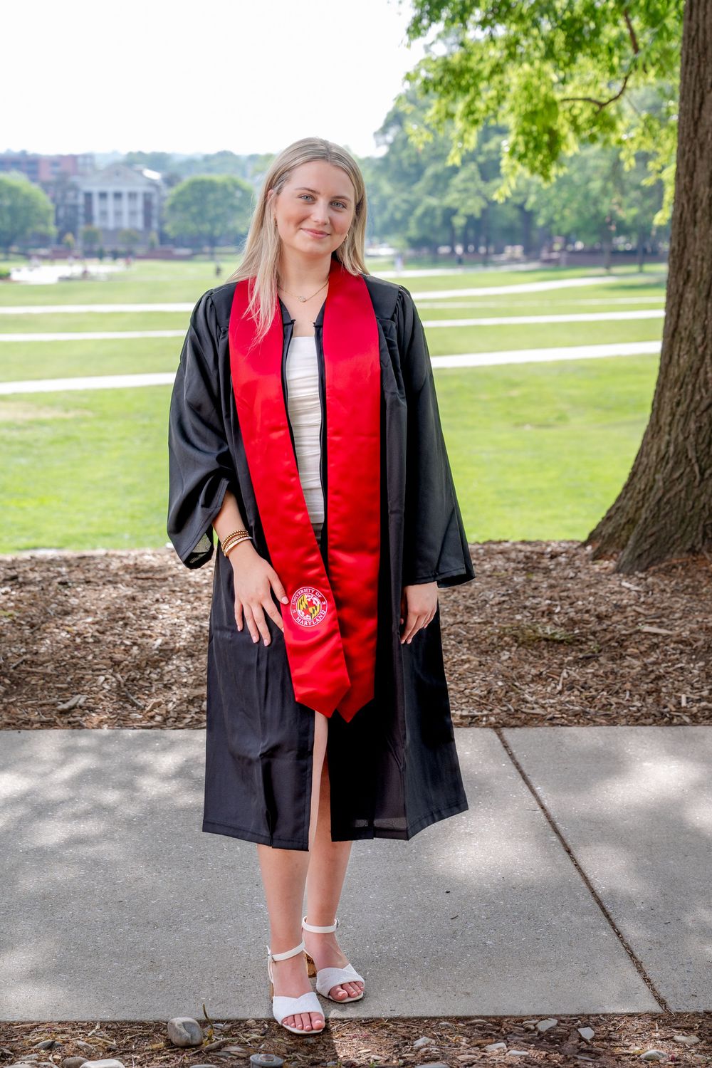 Graduate wearing black gown with red stole stands on UMD campus pathway.
