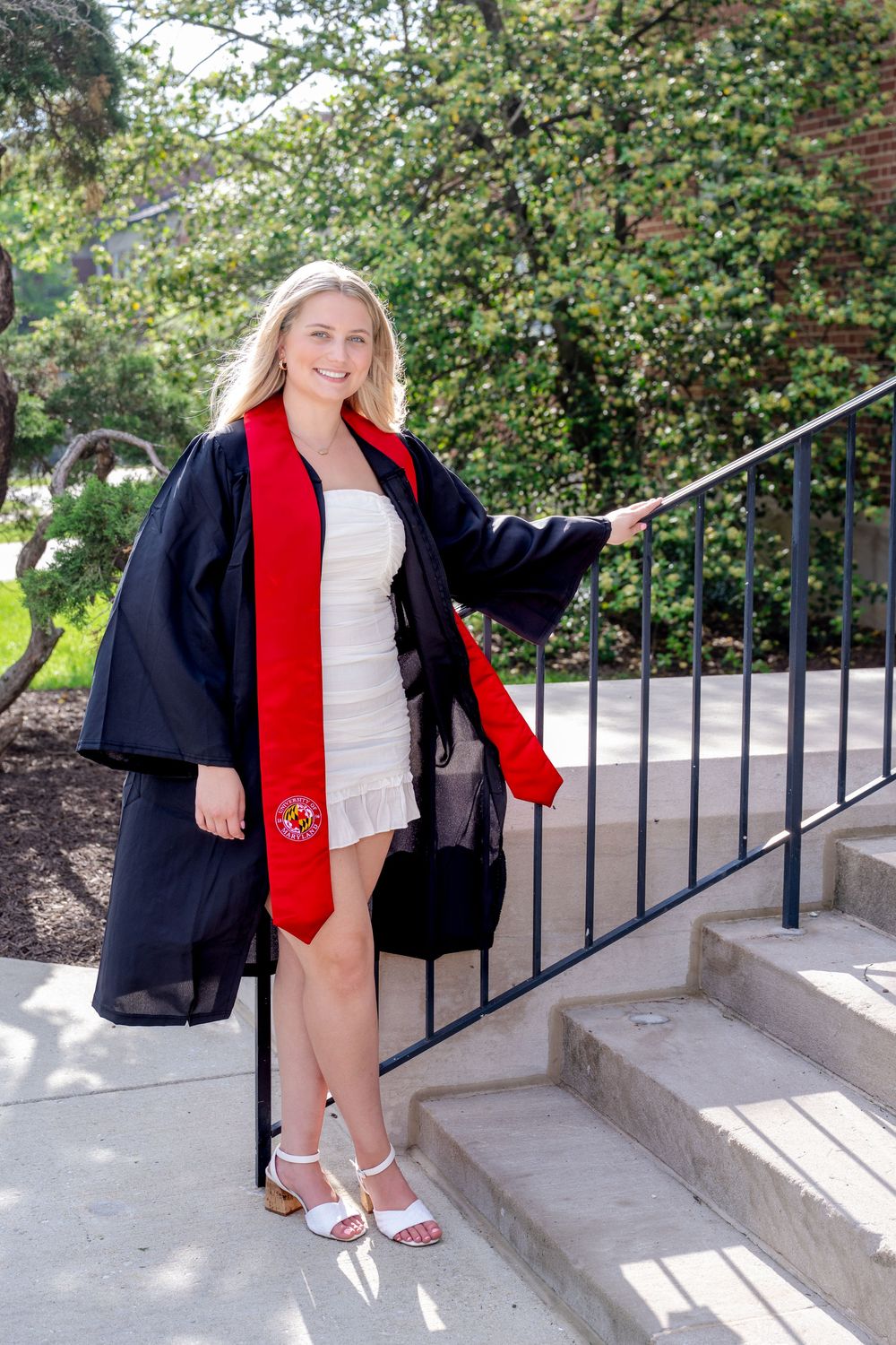 College graduate in a black graduation gown with red stole poses on concrete steps near greenery.