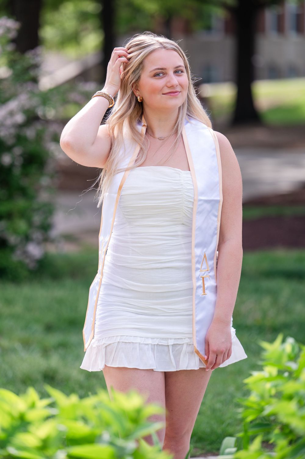 Outdoor photo of a college graduate wearing a white sleeveless summer dress with a ruched bodice and ruffle hem in a garden.