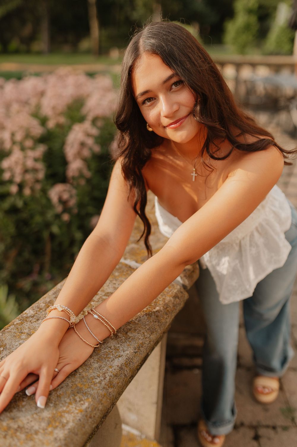 Summer portrait with delicate jewelry and white top leaning on stone wall with pink flowers.