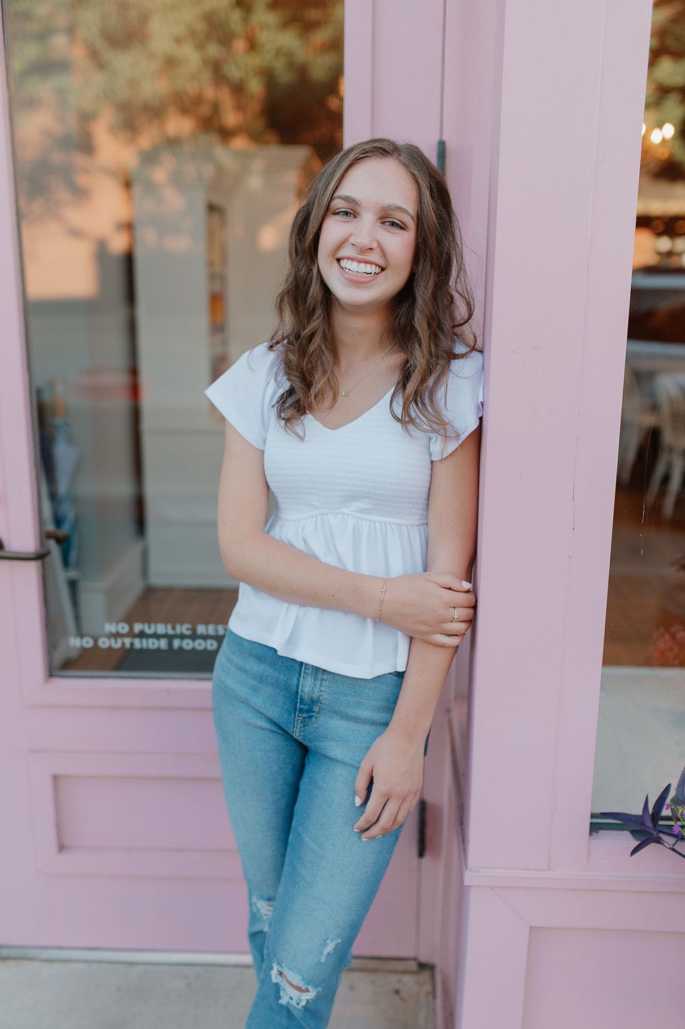 Standing by a pink door in a white peplum top and distressed jeans on a sunny day.