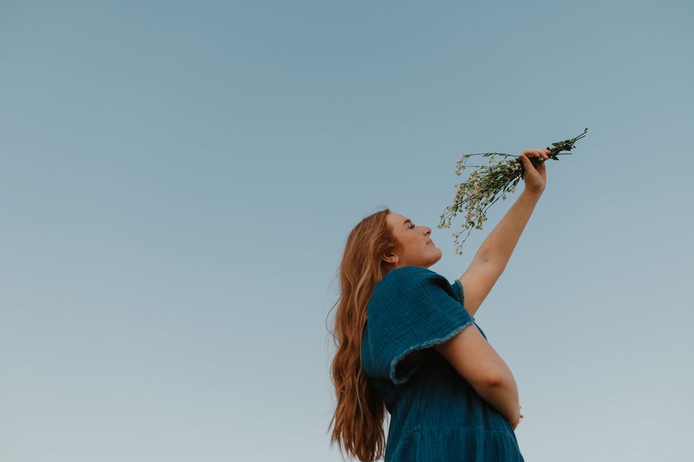 Long-haired person in blue dress holds up wildflowers against a bright summer sky.