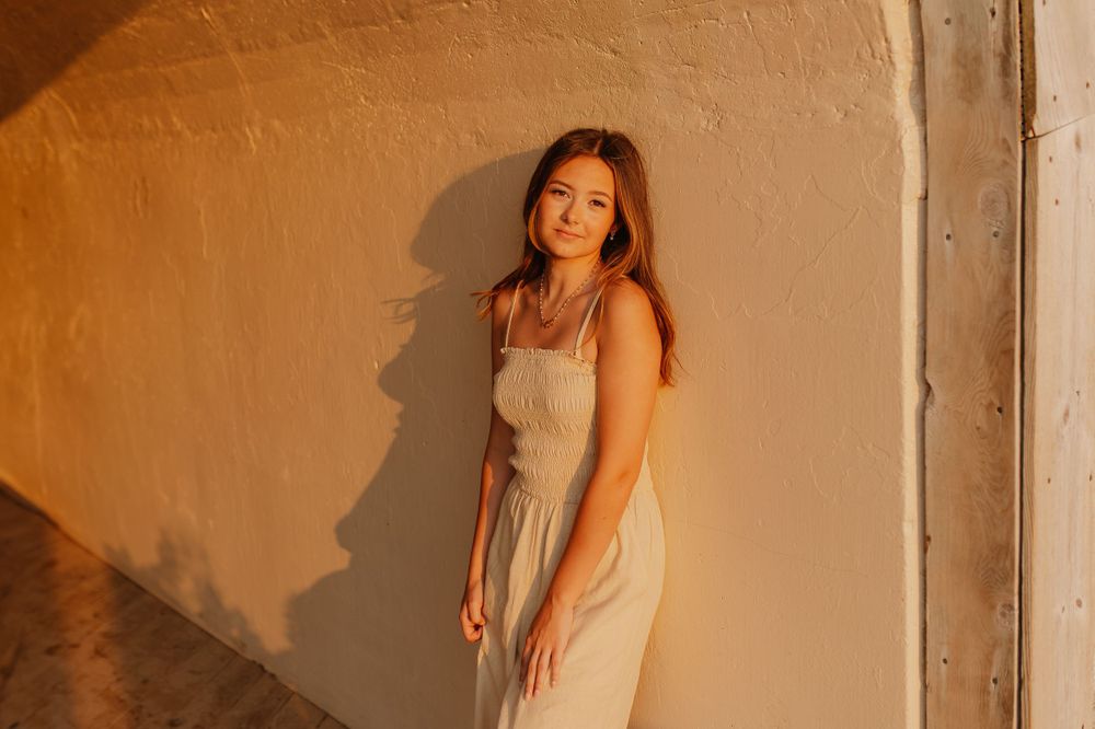 Woman in light dress poses against warm-toned wall with dramatic shadows.