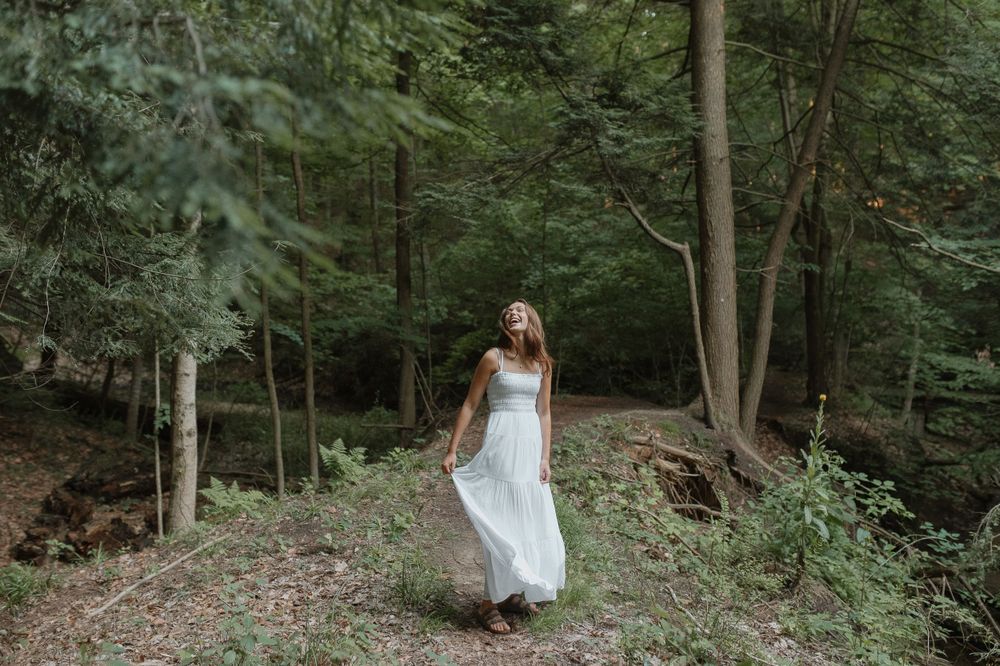 A white dress flows in the breeze as someone walks through a lush green forest.