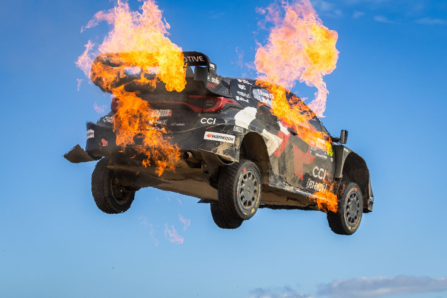 A dramatic sequence of a race car flipping and bursting into flames during a high-speed crash on a sunny day.