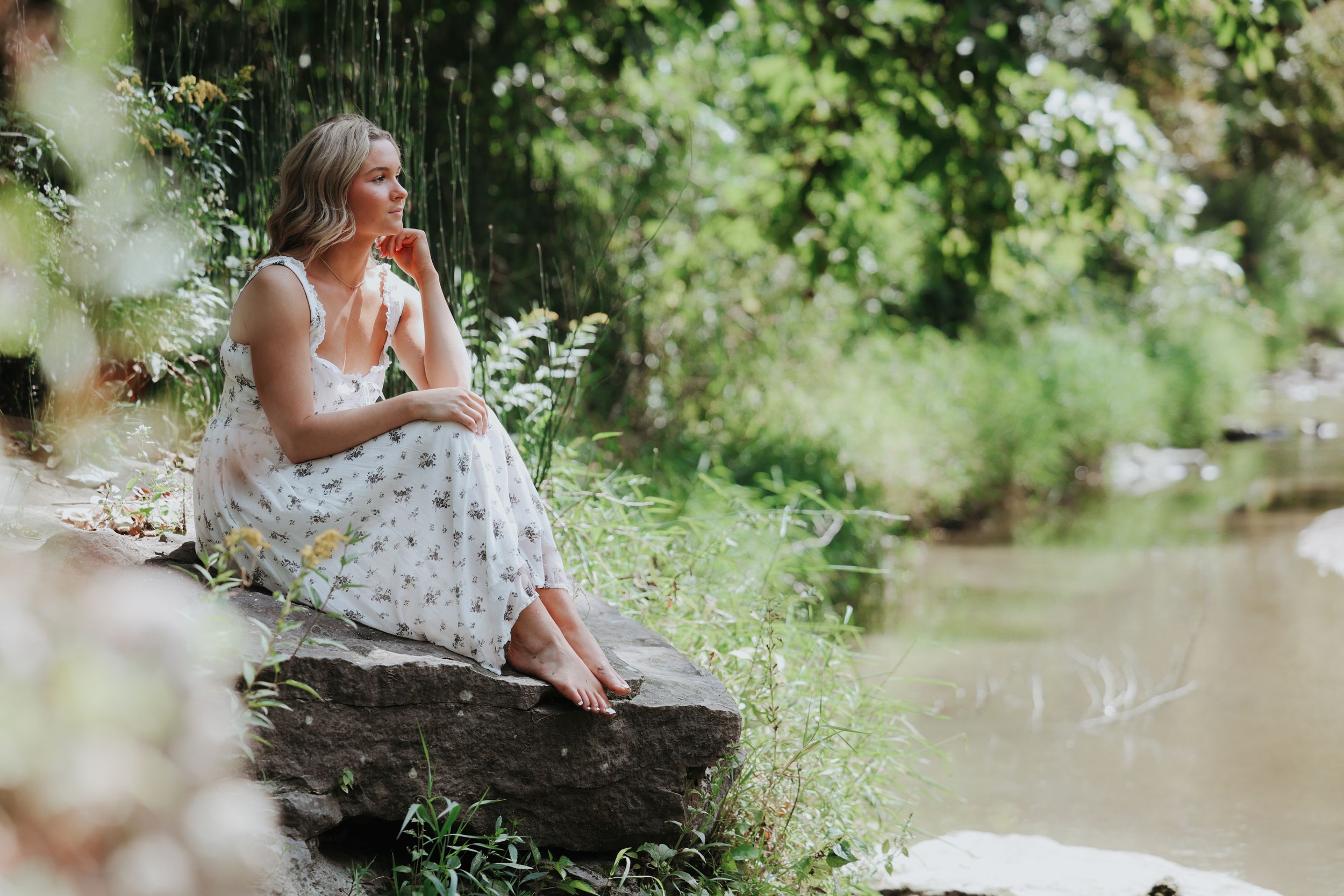 Senior photos, Allie is posing on a rock in front of a creek. The scene is very pretty, green, and soft.