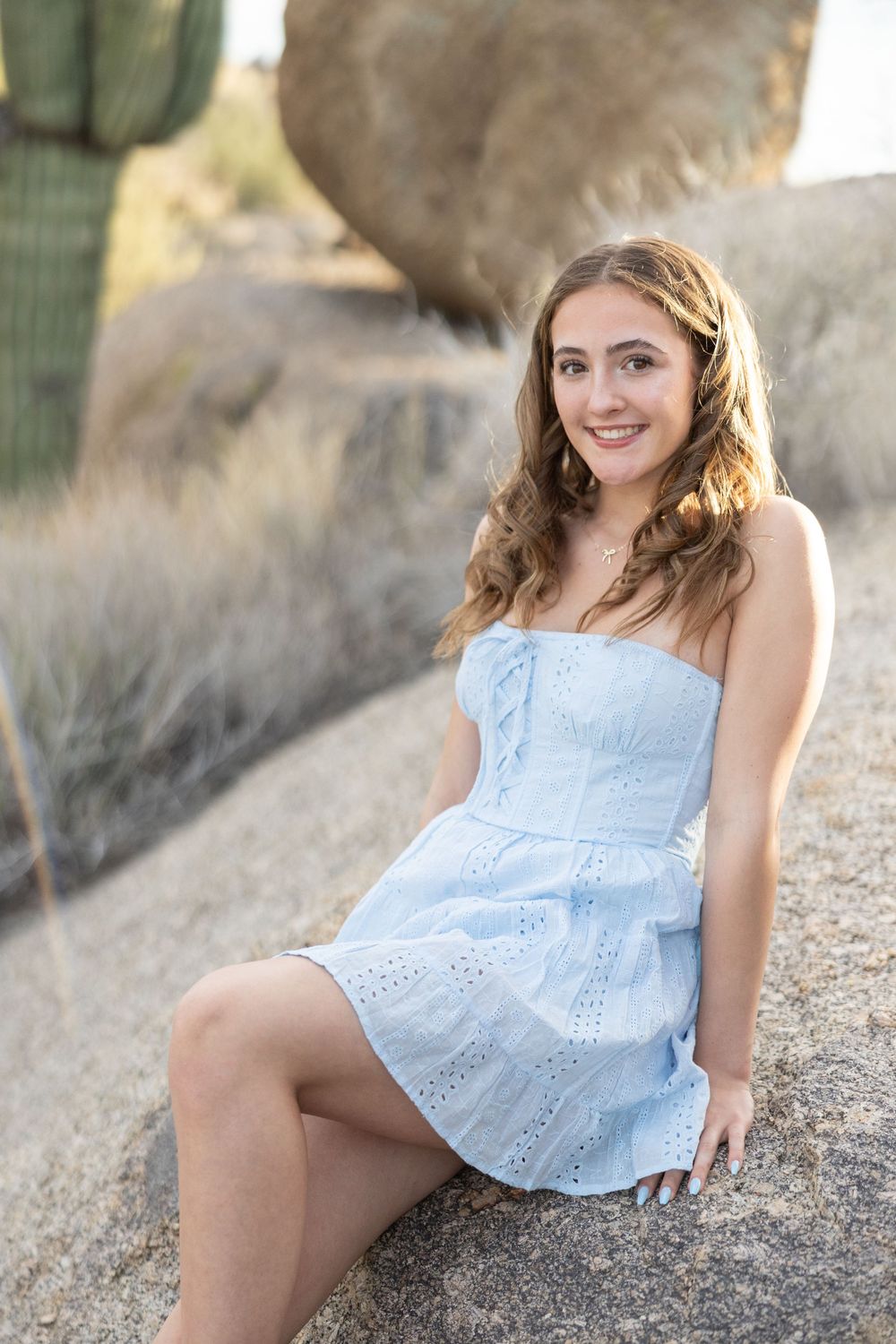 A person in a light blue eyelet dress sits casually on outdoor rocks during a sunny portrait session.