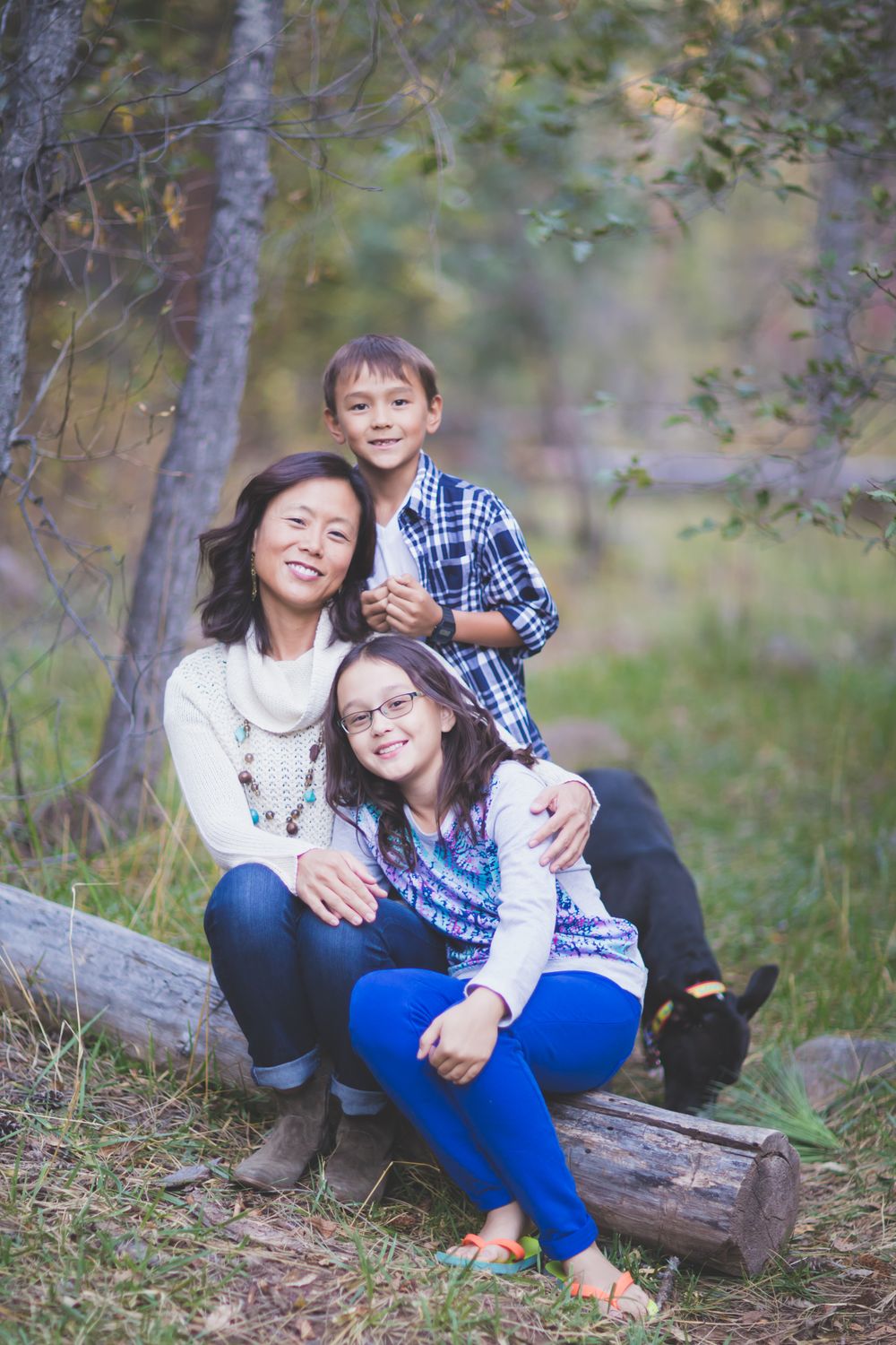 Family enjoying a casual outdoor photo session while sitting on a fallen log in a wooded forest setting.