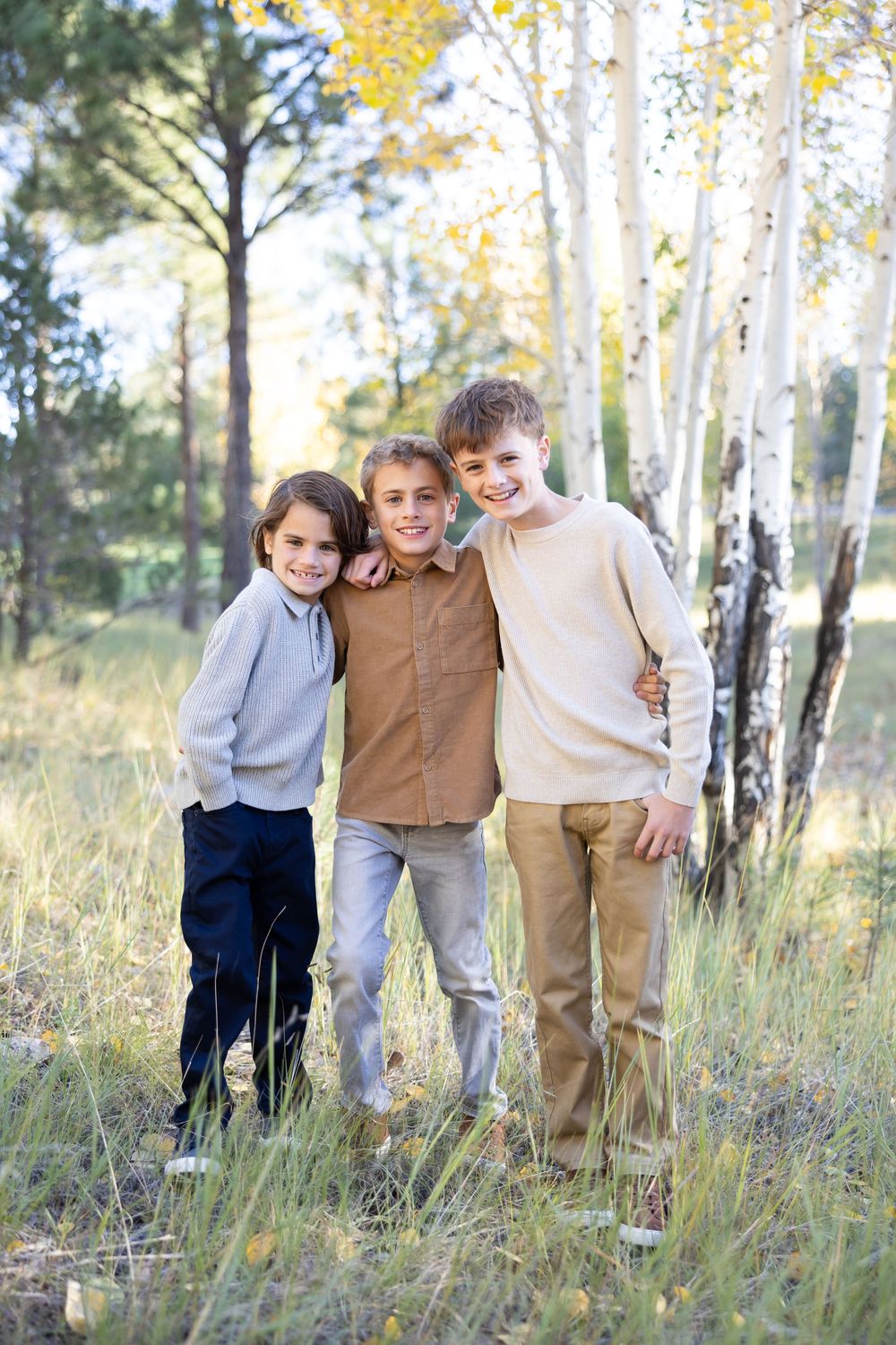 Three young boys pose together in casual clothing among aspen trees on a sunny autumn day.