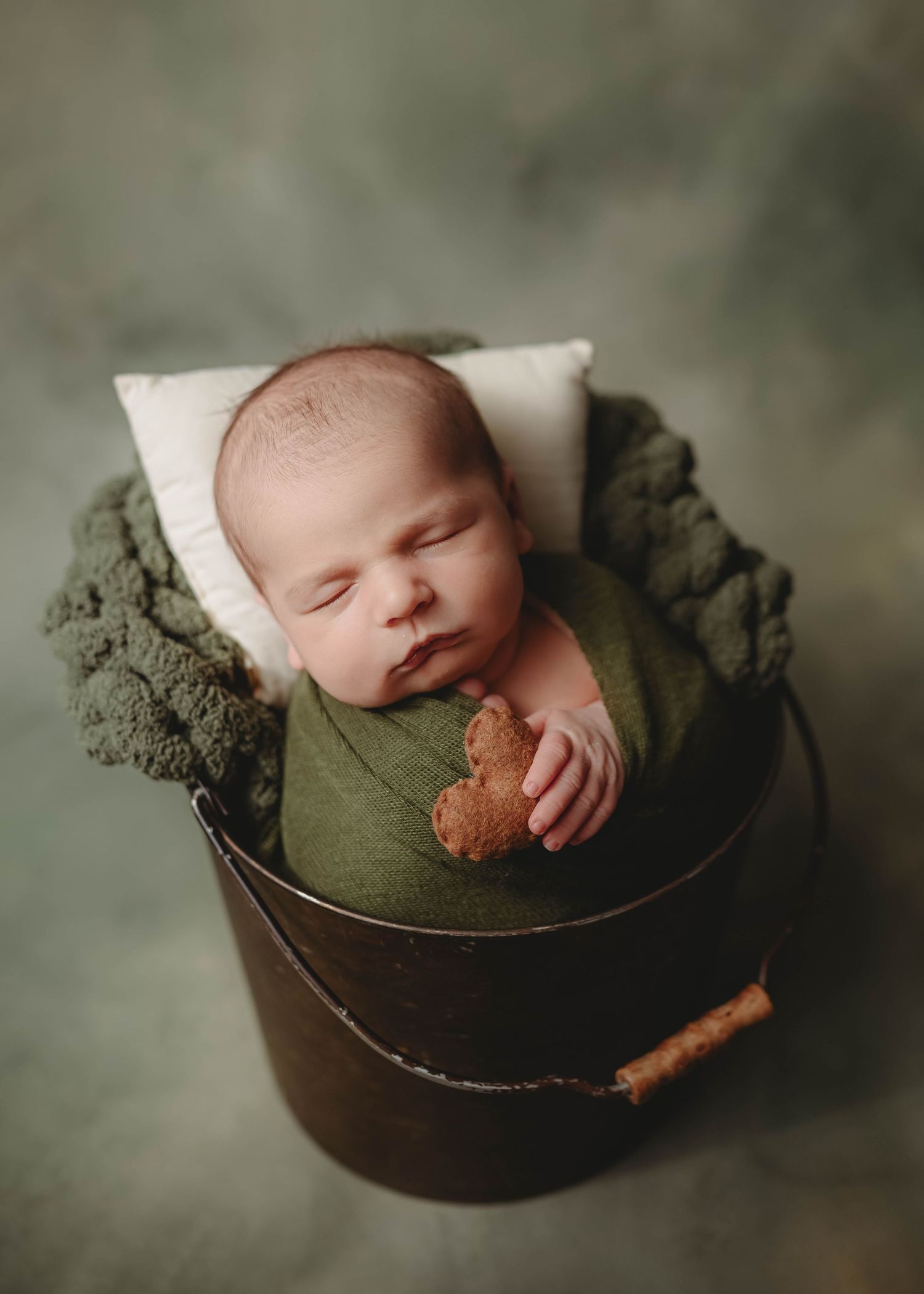 Sleeping newborn in olive green wrap nestled in rustic metal container.