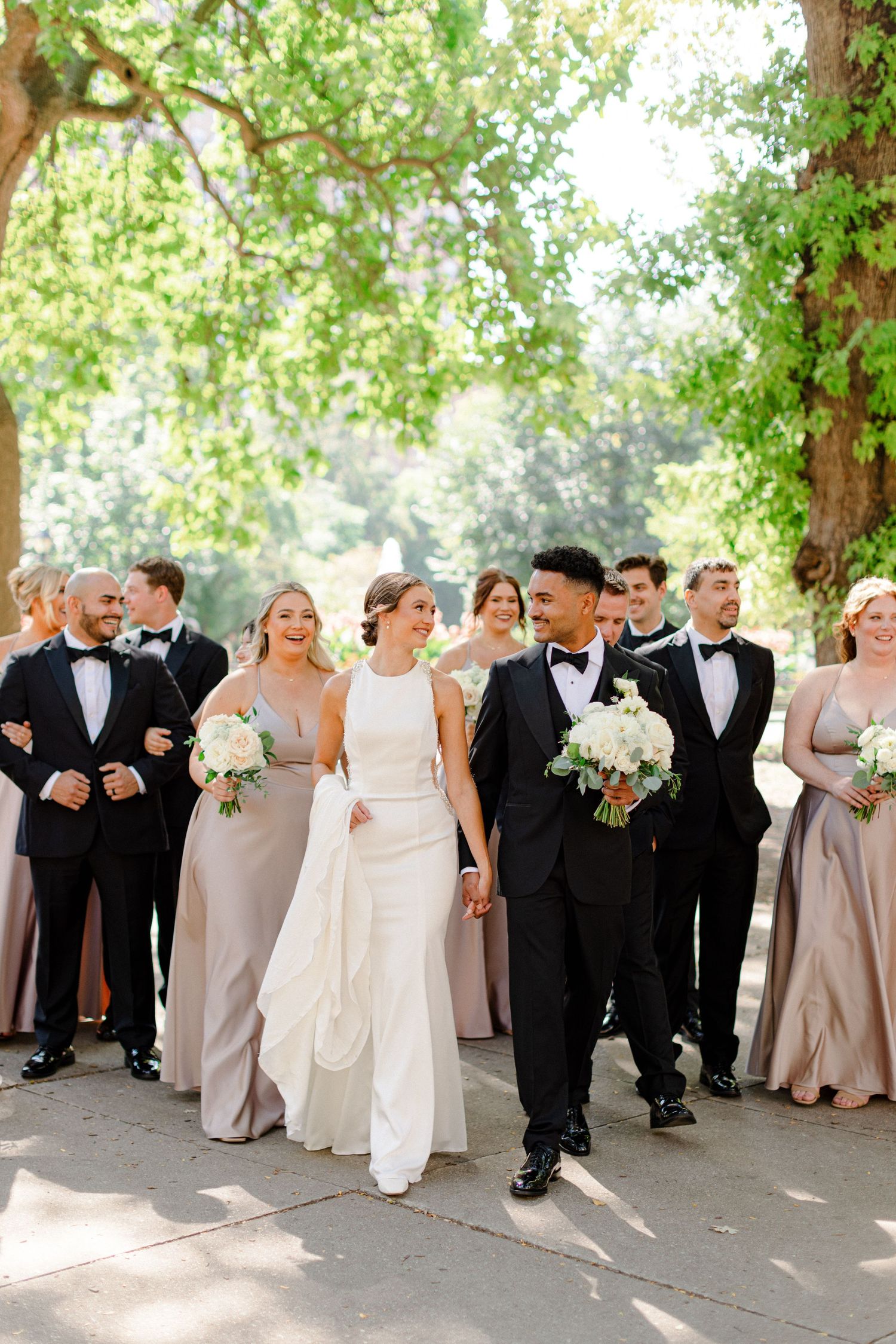 Wedding party walks together outdoors wearing formal attire in neutral tones under tree-lined path.