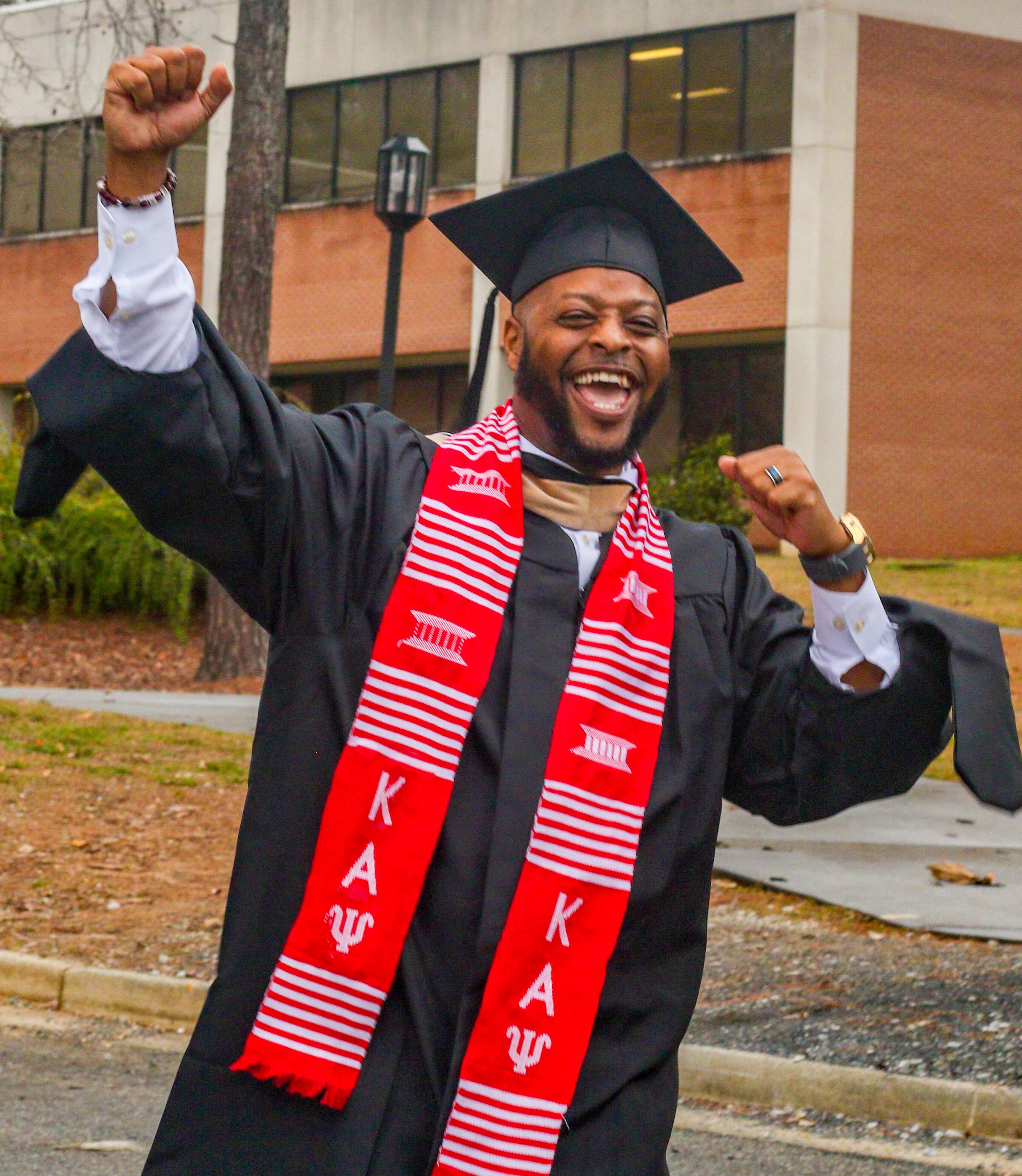Graduate in black cap and gown with red stole celebrates enthusiastically outside campus building.