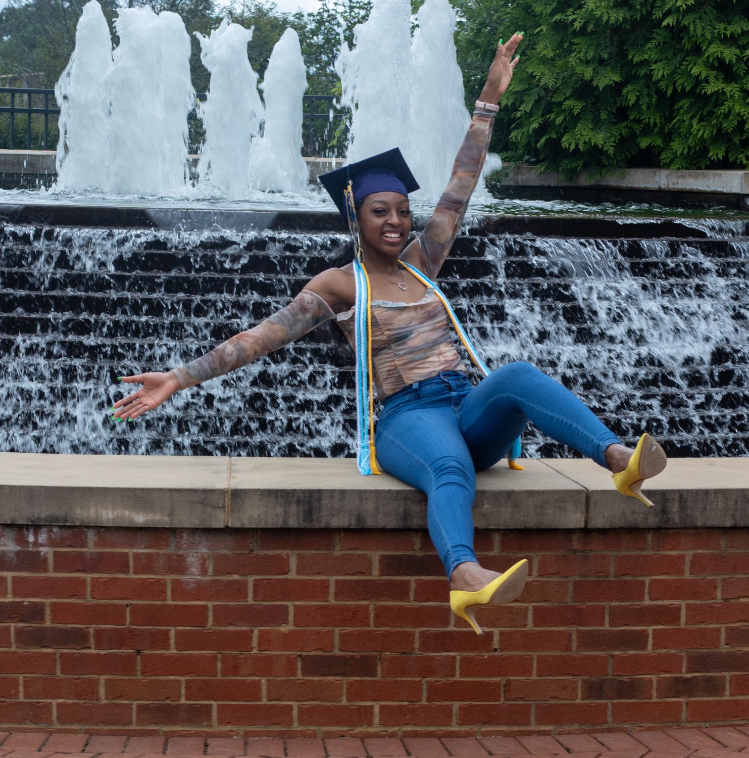 Graduate in cap and gown poses playfully by fountain during graduation celebration.
