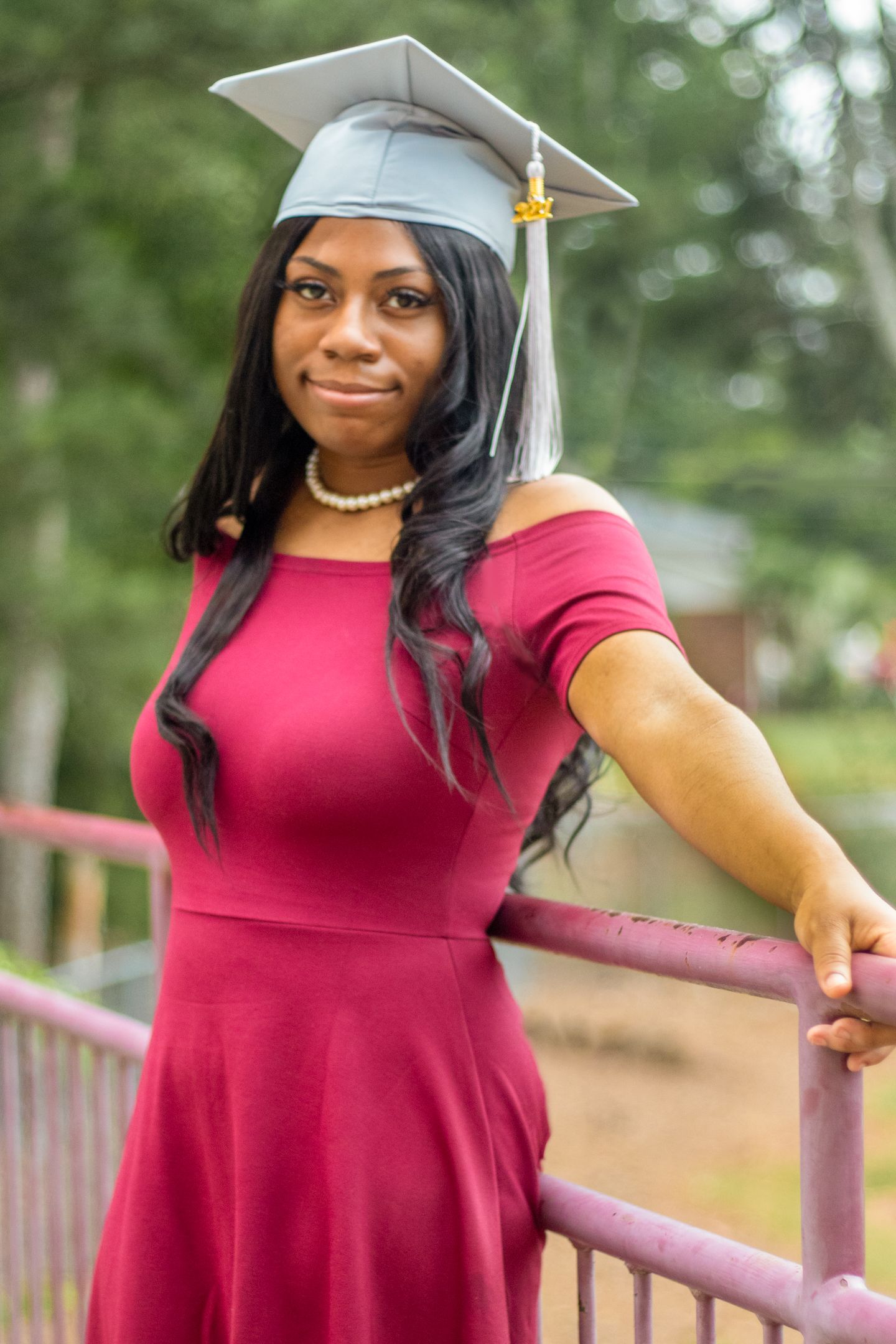 Graduate in red dress and silver cap poses against outdoor railing during graduation photoshoot.
