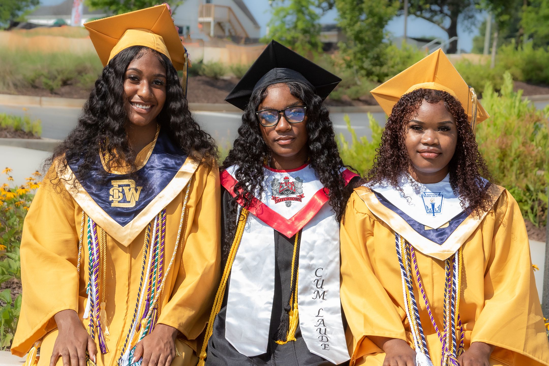 Three graduates in gold and navy academic regalia posing together on sunny campus grounds.