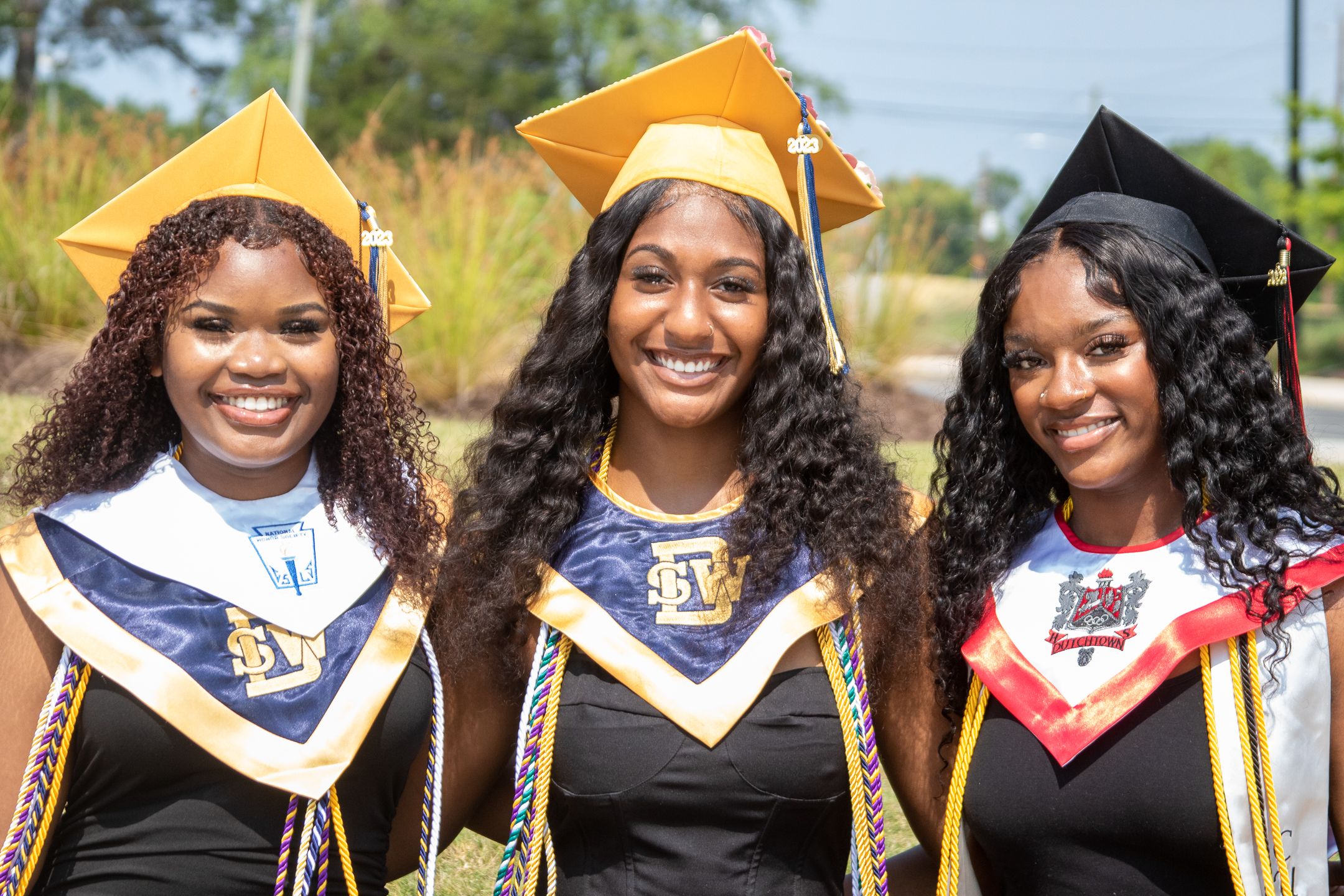Three graduates in academic regalia pose together outdoors at a commencement ceremony.