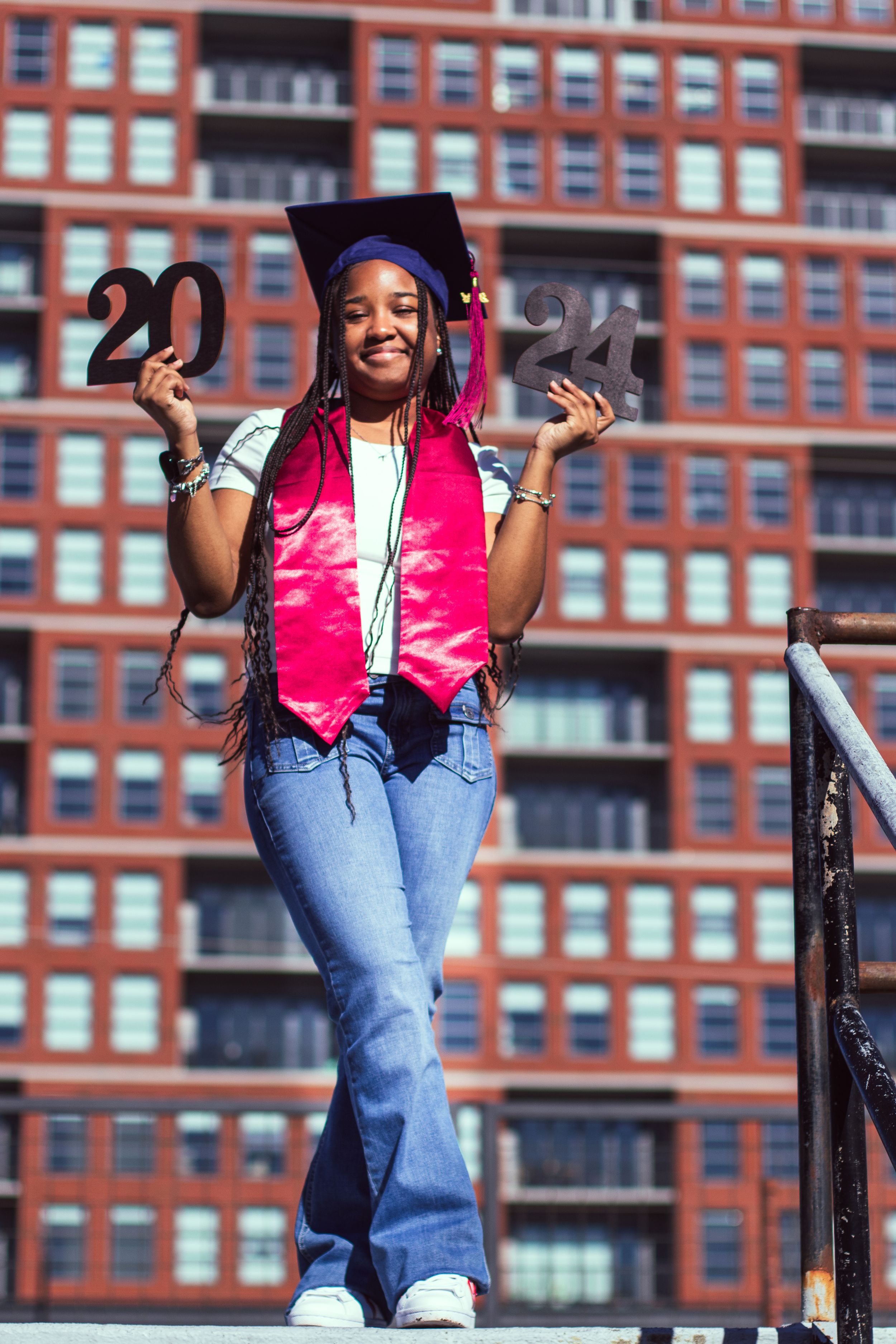 Graduate in pink stole holds up 2022 numbers against red brick building backdrop.