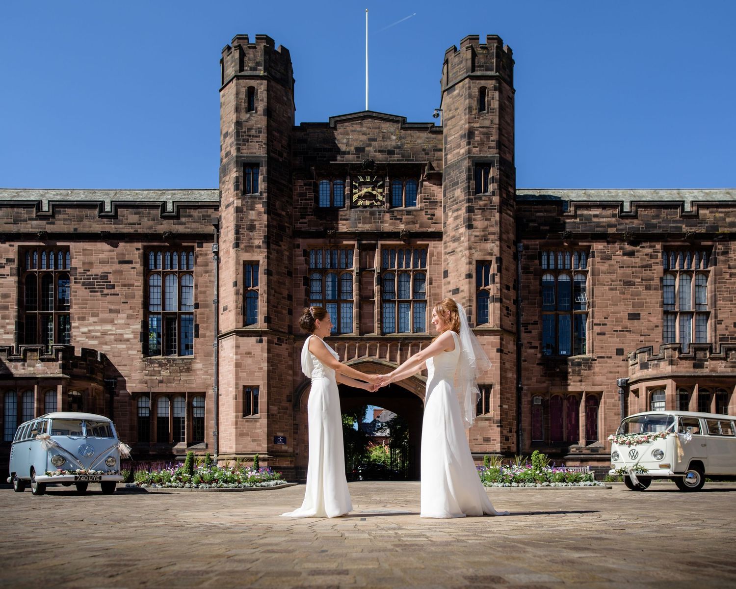 Two brides in white gowns hold hands outside a grand red sandstone castle with vintage VW campervans on a sunny day.