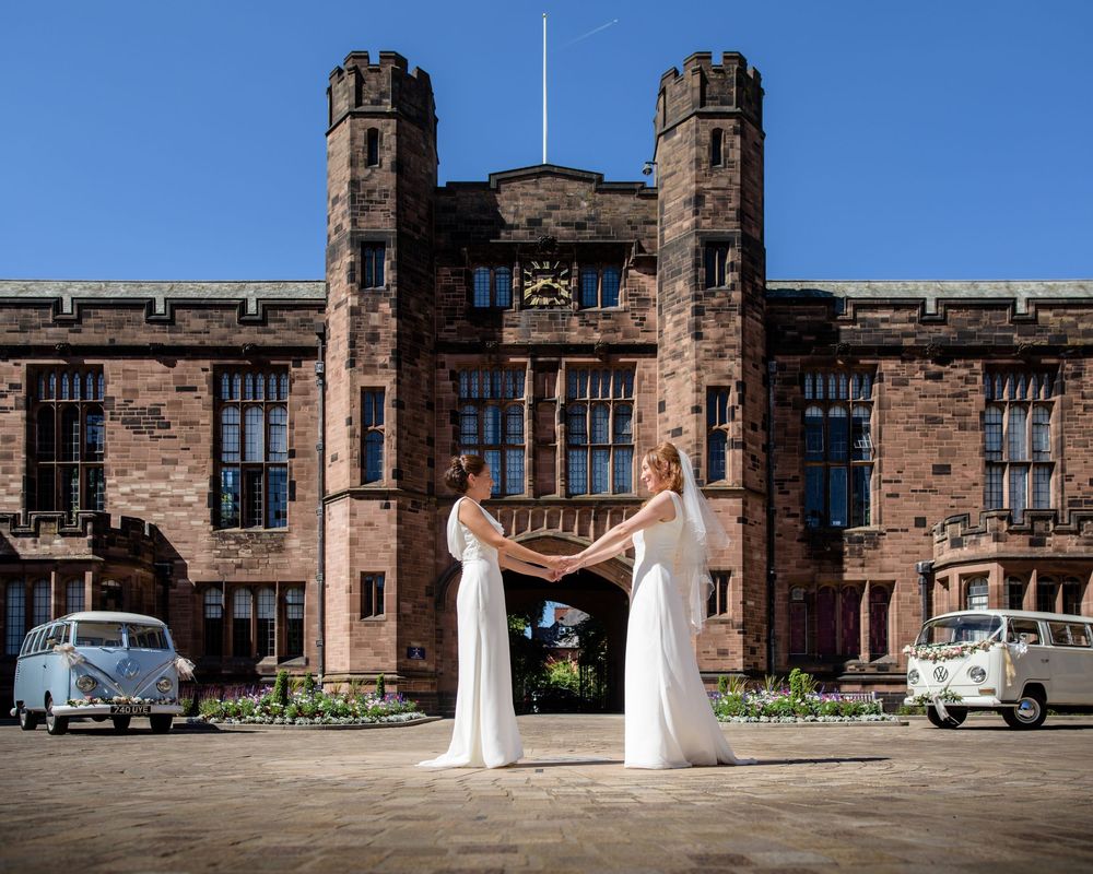 Two brides in white gowns hold hands outside a grand red sandstone castle with vintage VW campervans on a sunny day.
