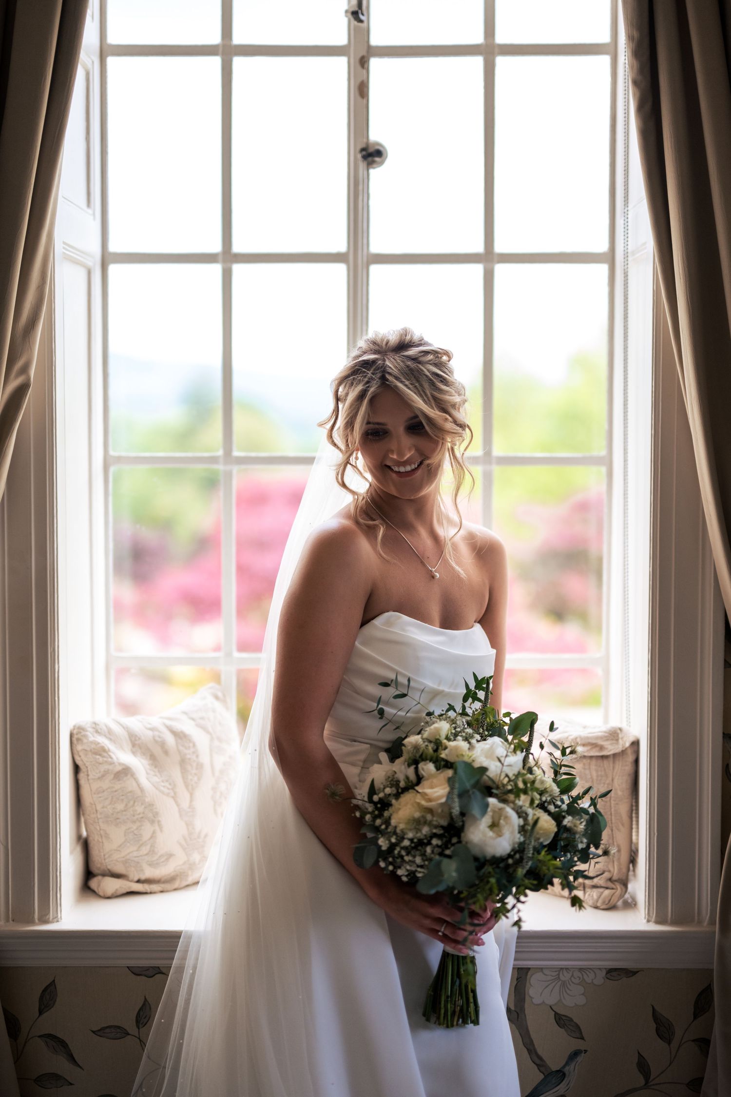 A bride in a white wedding dress holds a bouquet while standing near large windows in natural light.