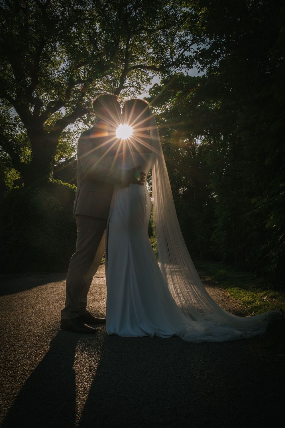 Silhouetted couple embraces in a romantic forest setting with sunburst filtering through trees at sunset.