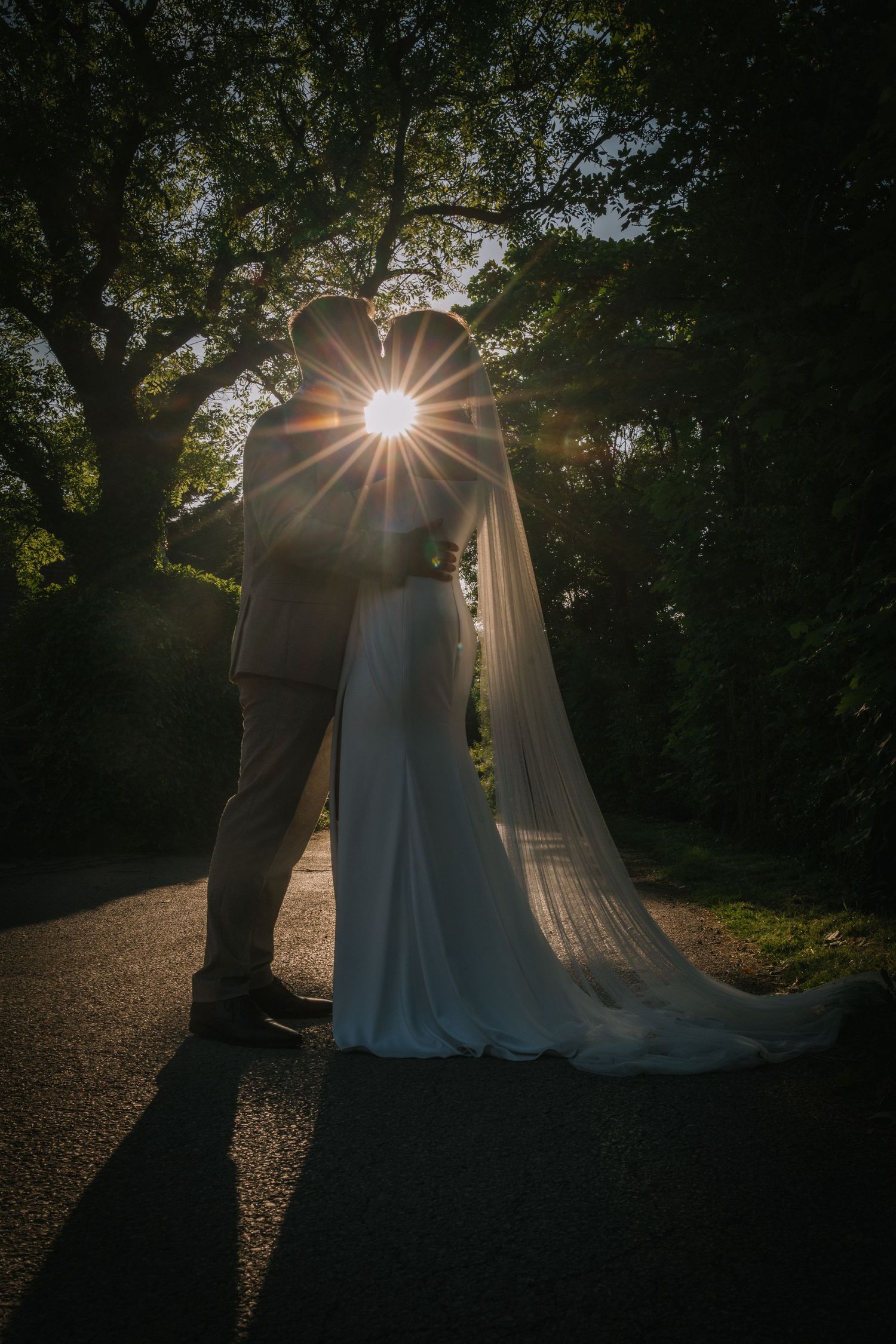 Silhouetted couple embraces in a romantic forest setting with sunburst filtering through trees at sunset.