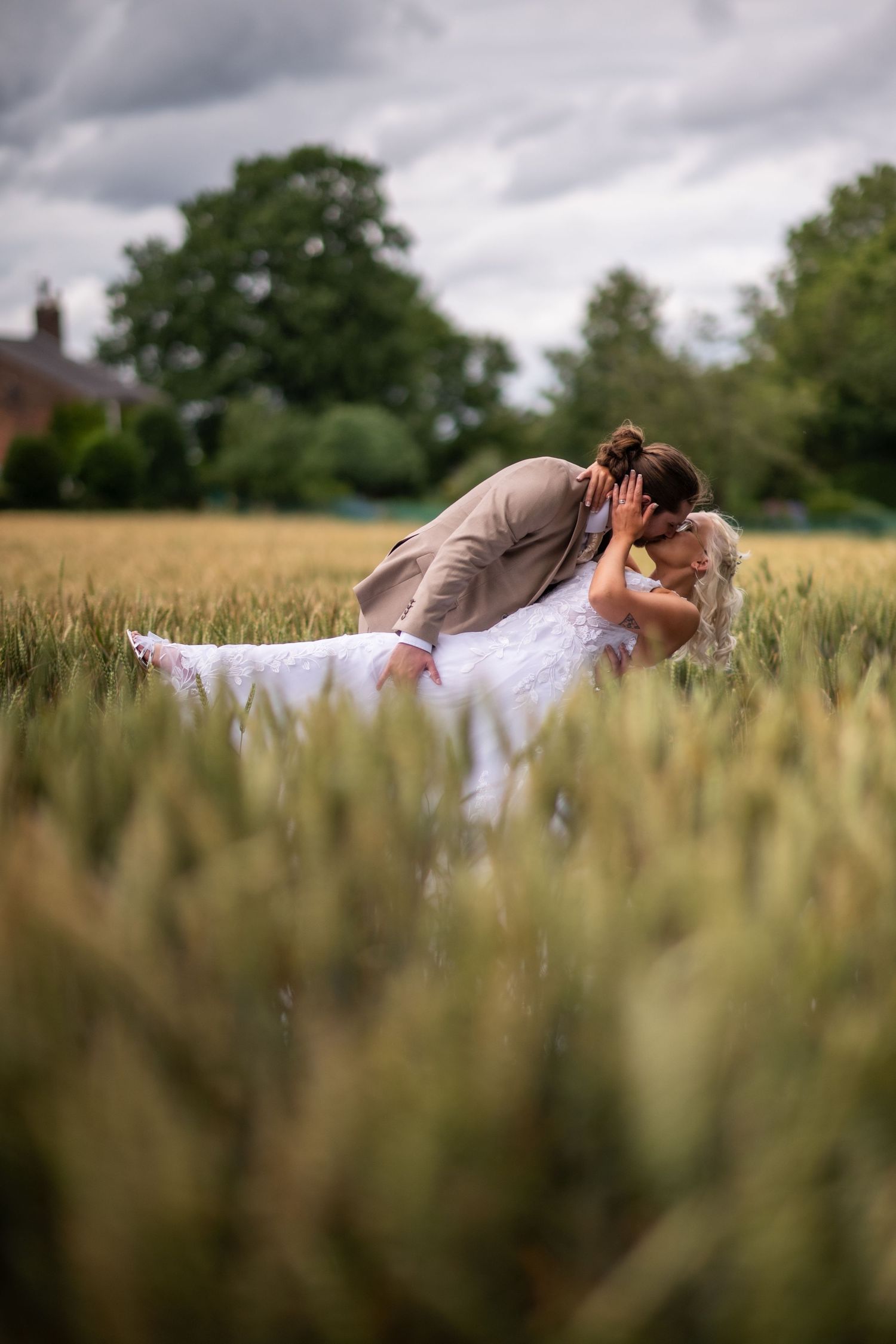 A romantic couple in wedding attire lies together in a golden wheat field under dramatic cloudy skies.