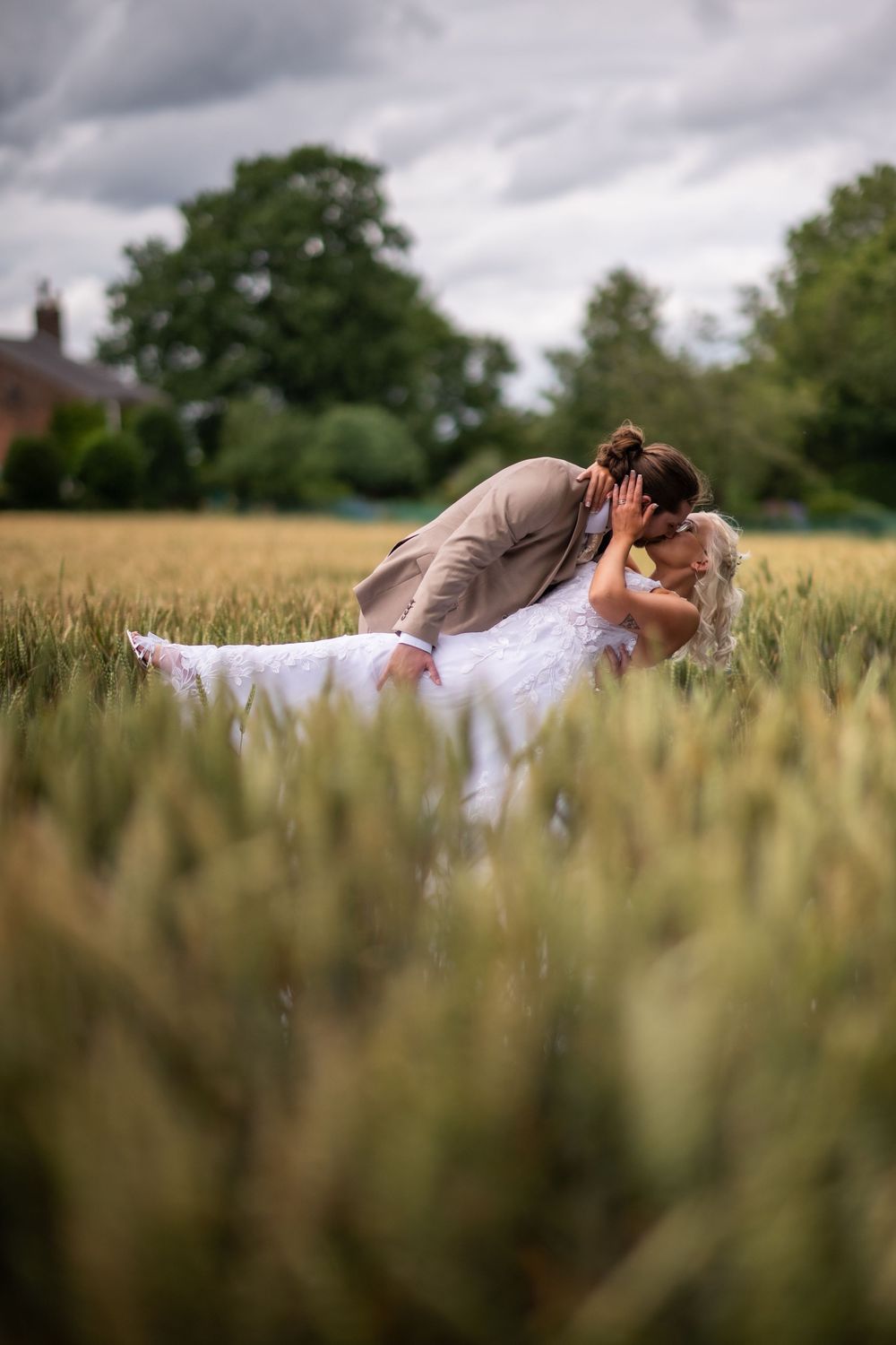 A romantic couple in wedding attire lies together in a golden wheat field under dramatic cloudy skies.