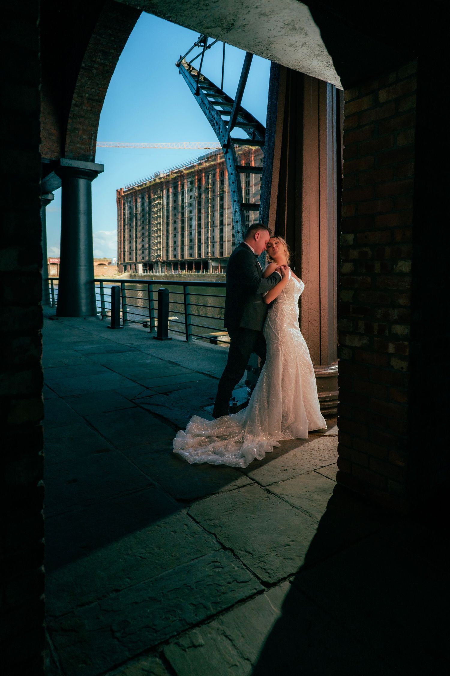 Bride and groom share a romantic kiss framed by a stone archway with a waterfront bridge and building backdrop.
