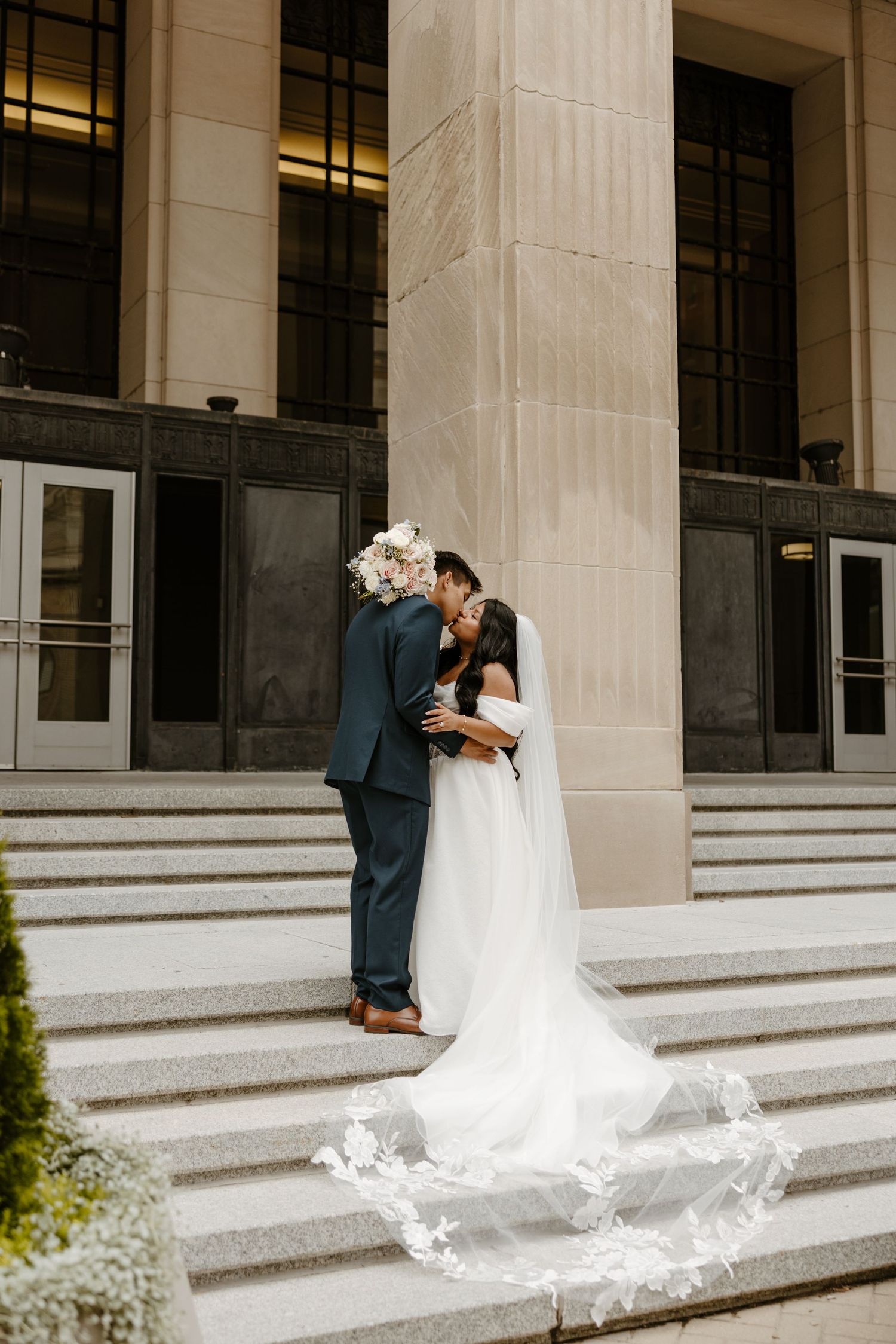 Bride and groom share a kiss on grand building steps, her lace veil trailing elegantly, holding a white floral bouquet. DFW photographer