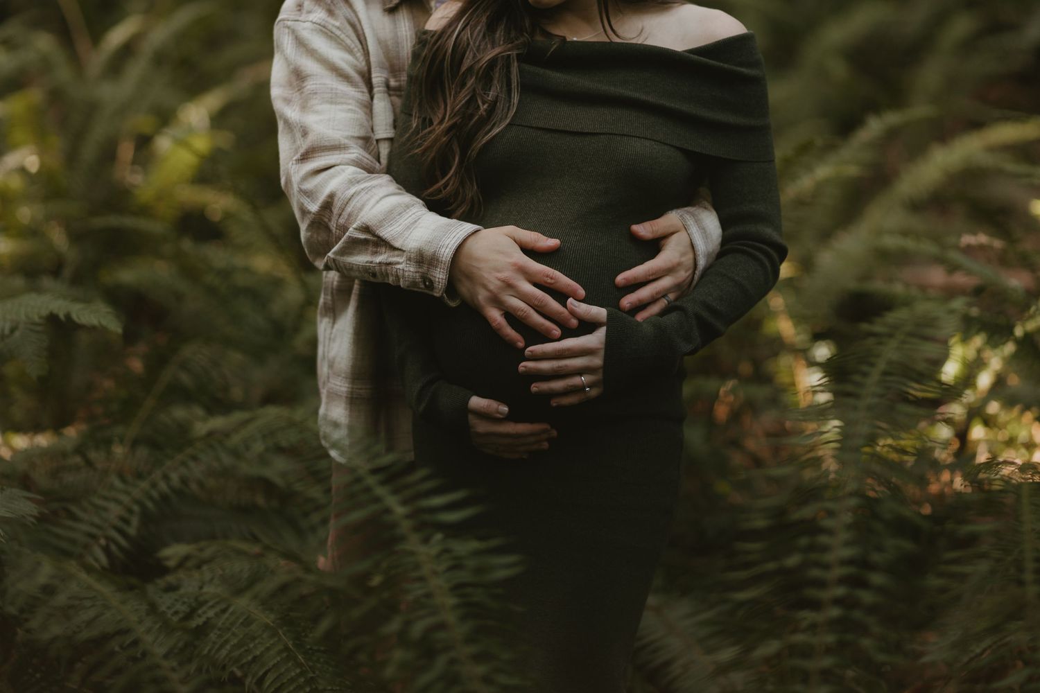 Silhouetted hands tenderly hold a pregnant belly among ferns in moody lighting.
