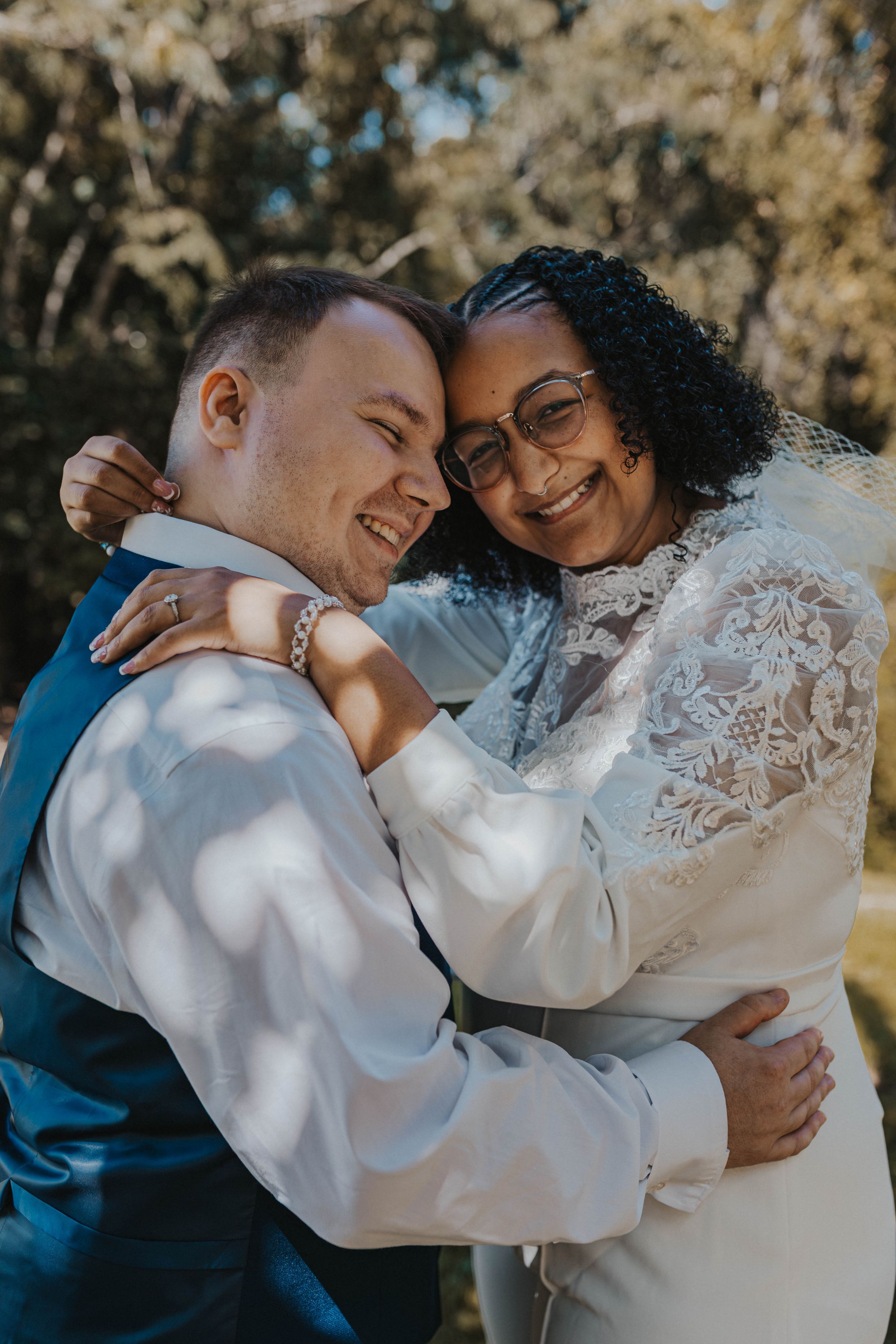 A joyful couple embraces outdoors on their wedding day, wearing white and blue attire in dappled sunlight.