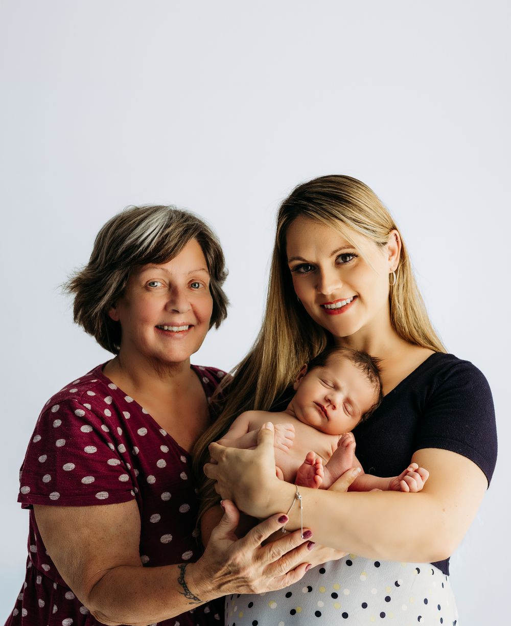 Three generations pose for a heartwarming studio portrait with a resting infant against a light gray backdrop. session in Oakton, VA