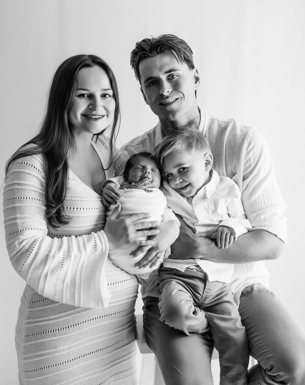 Black and white family photo series showing tender moments between parents and baby in matching white outfits.