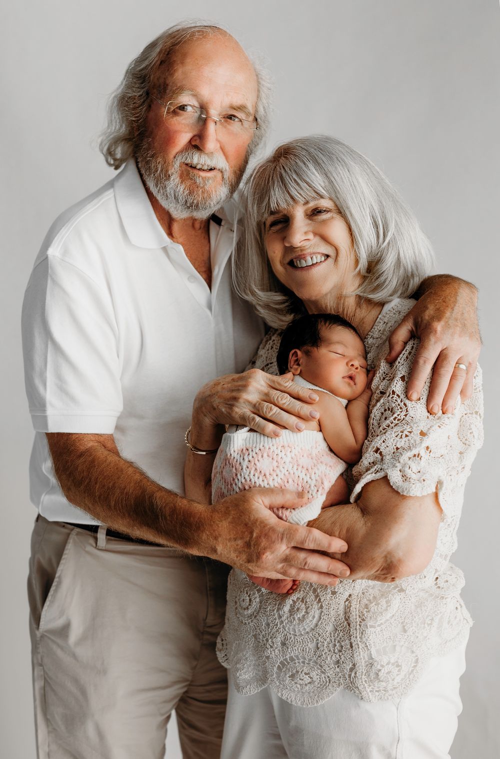 Elderly couple in white clothing holding and gazing lovingly at a sleeping newborn baby against a light background. session in Fairfax, VA