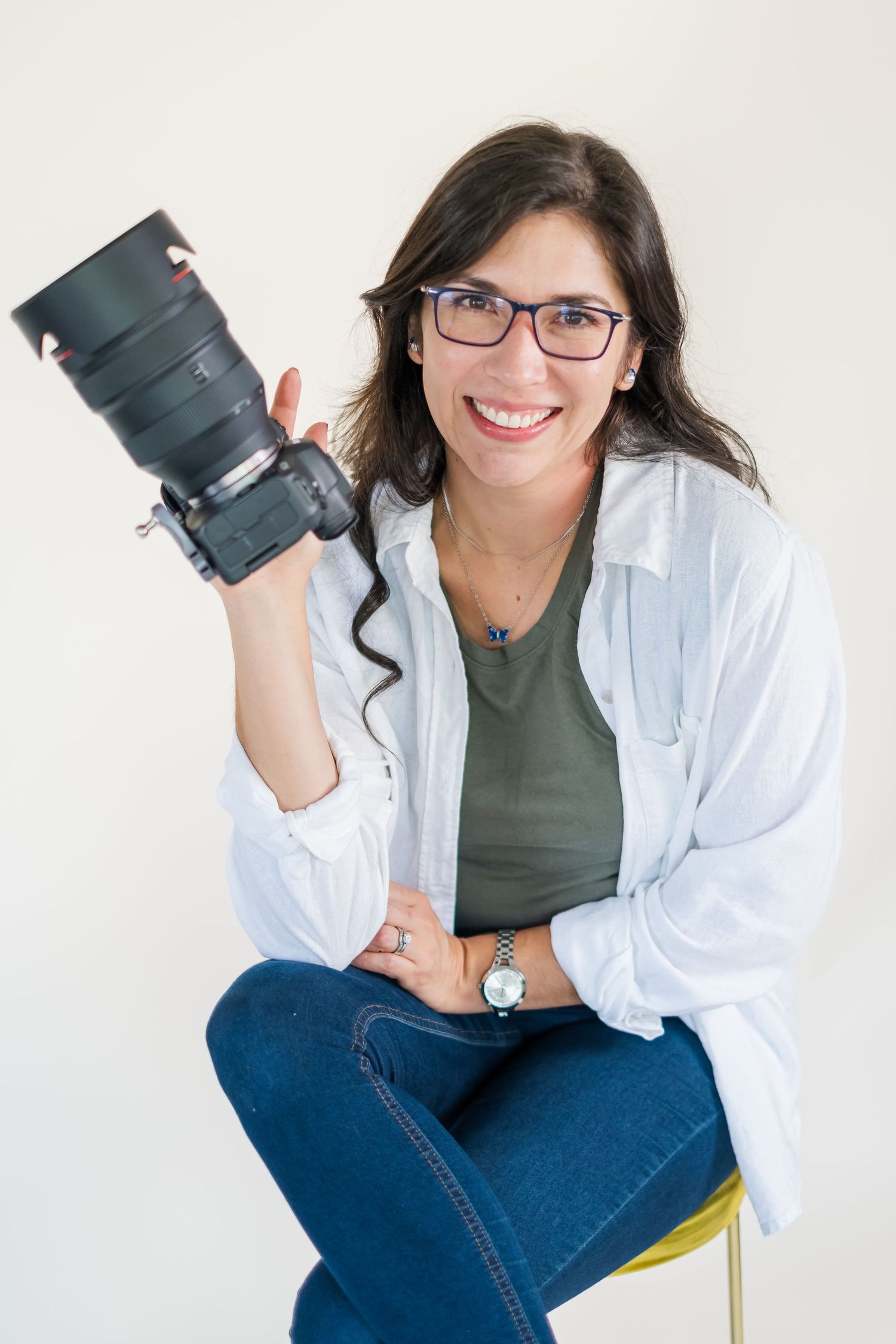Professional photographer in white button-down shirt and jeans poses with camera against white backdrop.