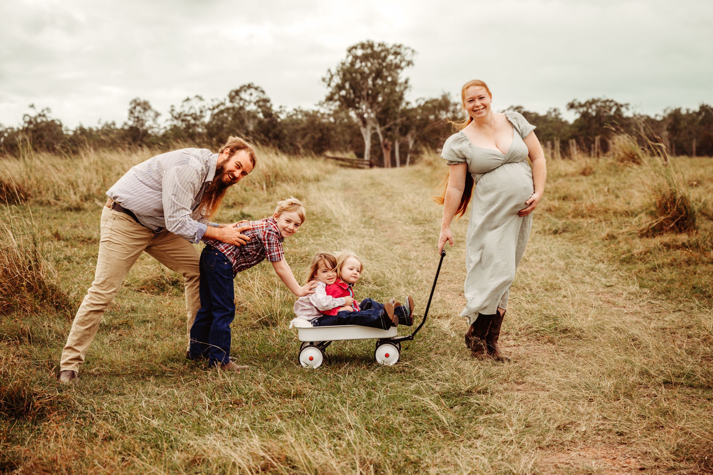 Pregnant woman pulls children in wagon while man pushes during outdoor family maternity photo shoot in grassy field.