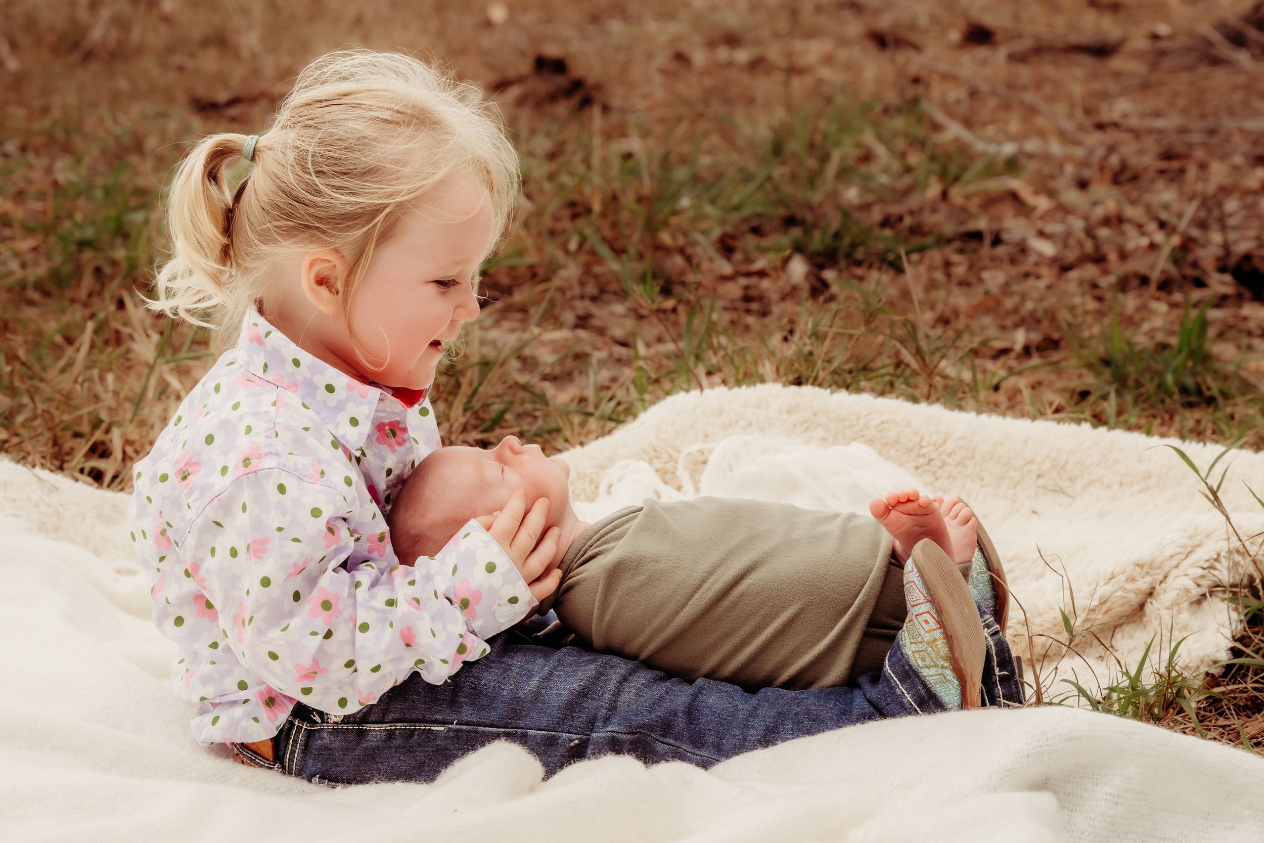 Young blonde girl in a polka dot shirt sits on a cream blanket outdoors, gently holding a baby doll in her lap on a grassy field.